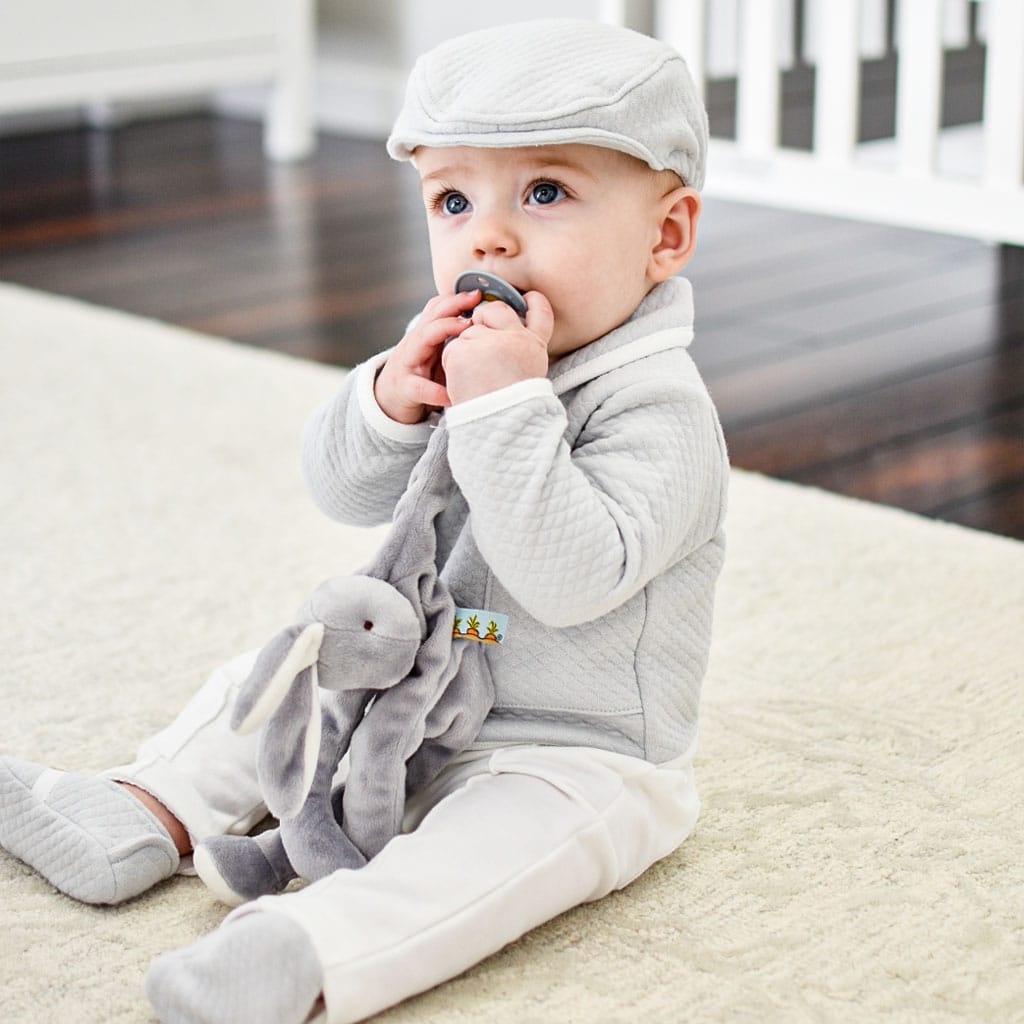 A baby dressed in a light gray outfit and cap sits on a cream-colored carpet, happily holding and chewing on the Grey Silly Bunny Buddy, which doubles as a pacifier holder. Nearby, the charm of the scene is enhanced by the dangling pacifier clip. The setting is completed with wooden flooring and a white crib in the background, making it an ideal backdrop for showcasing a perfect baby shower gift.