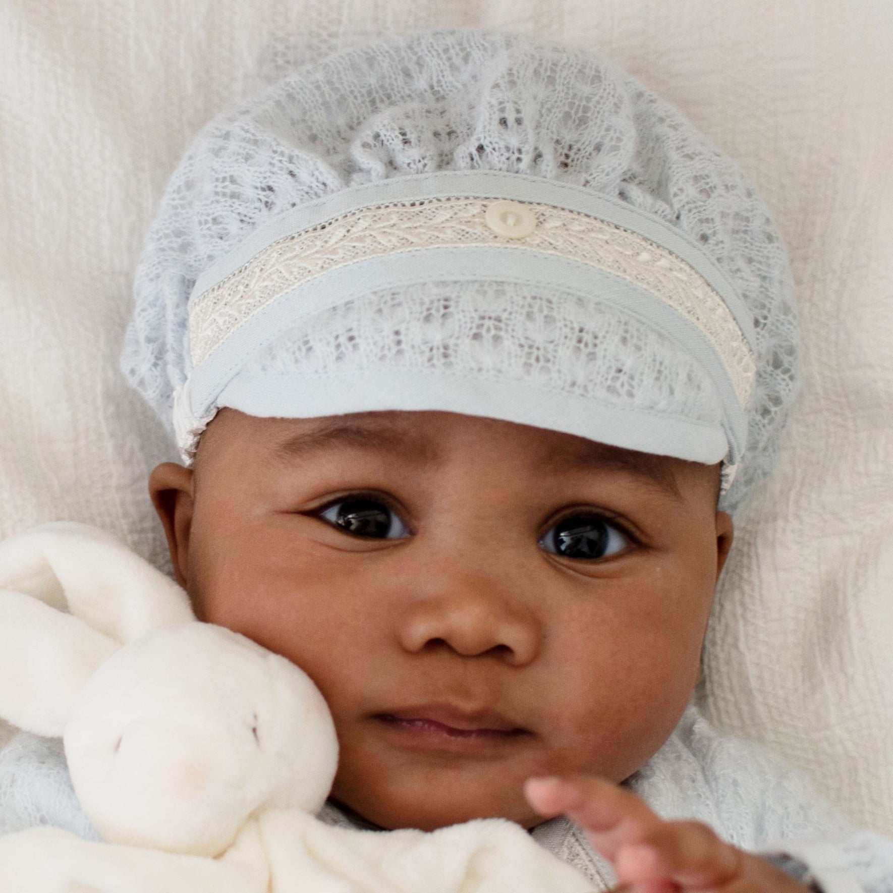 Infant cuddled with a plush bunny, wearing the Harrison Blue Knit Christening Hat with intricate lace and ivory accents.