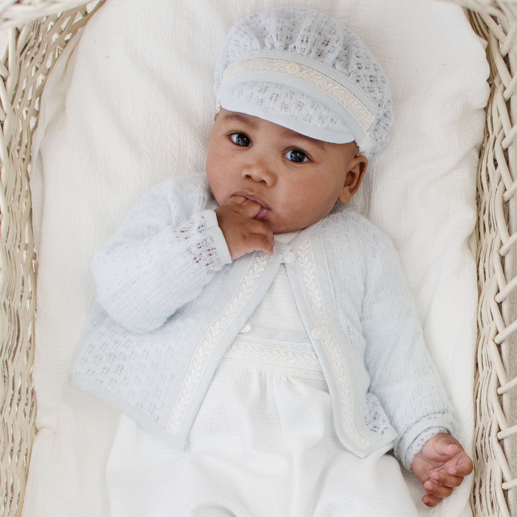 Baby in a cradle dressed in a blue knit christening sweater and coordinating white outfit, showcasing a serene look.