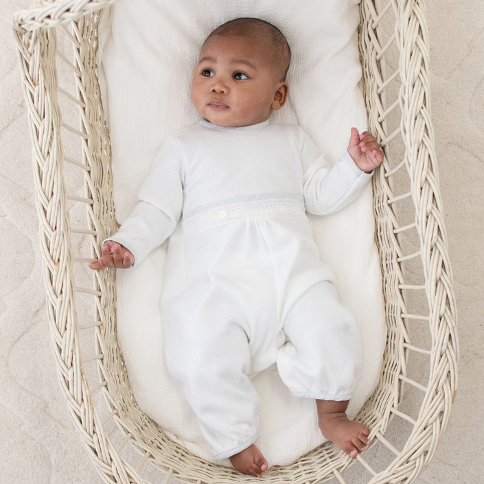 Infant dressed in the Harrison Long Sleeve Christening Romper, lying peacefully in a basket with intricate lacework and soft white design.