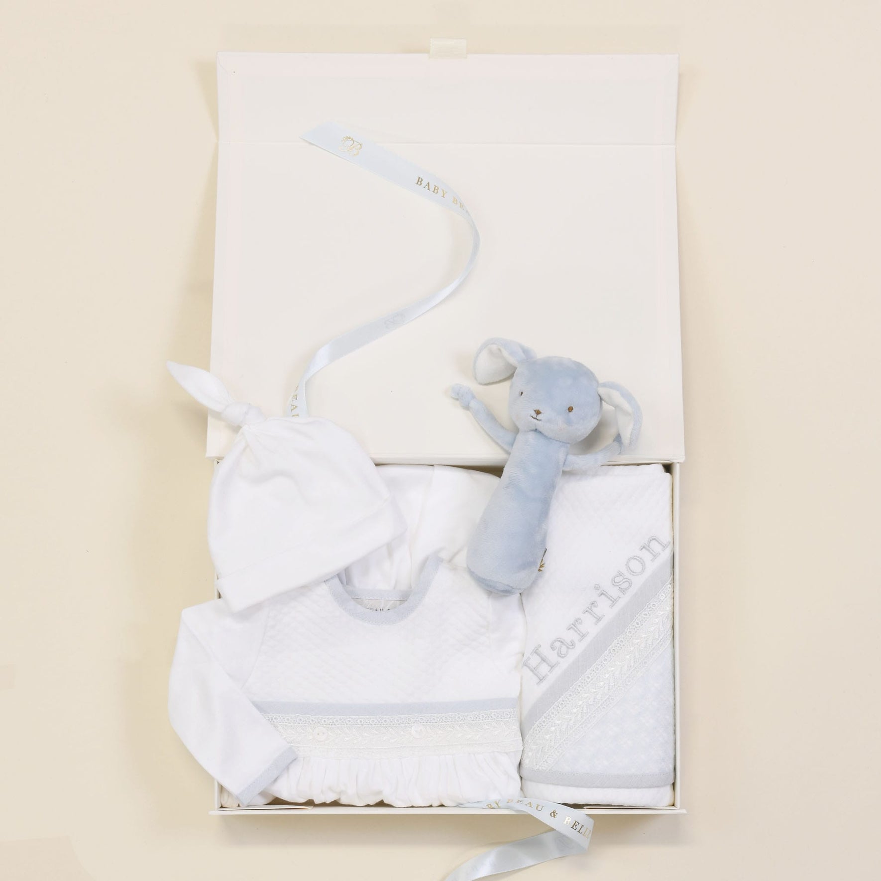 The Harrison Newborn Gift Set, available at a 10% discount, makes an ideal baby shower gift. This elegant white gift box includes a Pima cotton outfit set with a white hat and onesie, complemented by a cozy blue plush bunny toy. A personalized baby blanket embroidered with "Harrison" adds an extra touch of charm to this delightful ensemble.