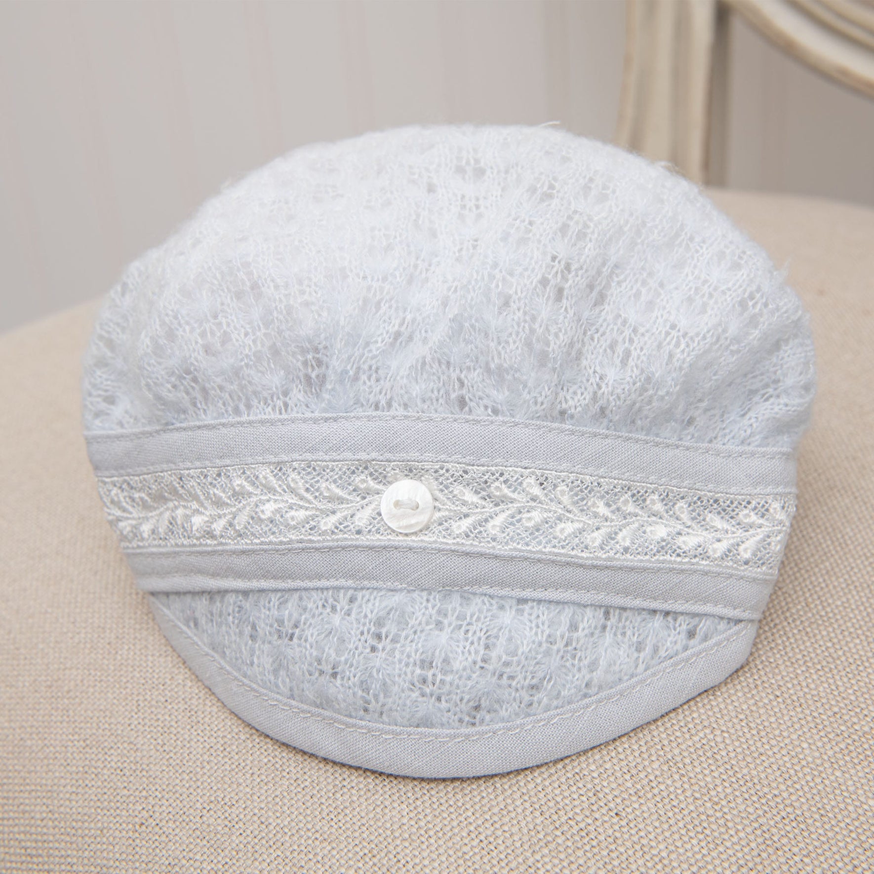 The Harrison Newborn Knit Hat, featuring a light gray color with a textured knit pattern, ivory Venice lace detailing, and a decorative button at the center front, is placed on a beige surface.
