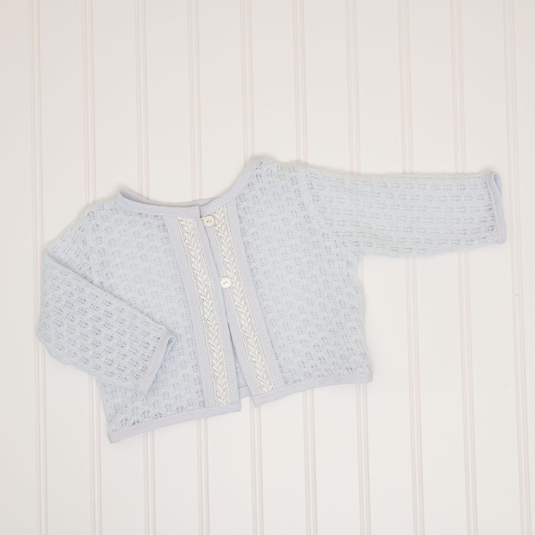 The Harrison Newborn Sweater, a soft blue knit sweater featuring a textured pattern and two buttons on the front, is laid flat on a white, subtly striped background.