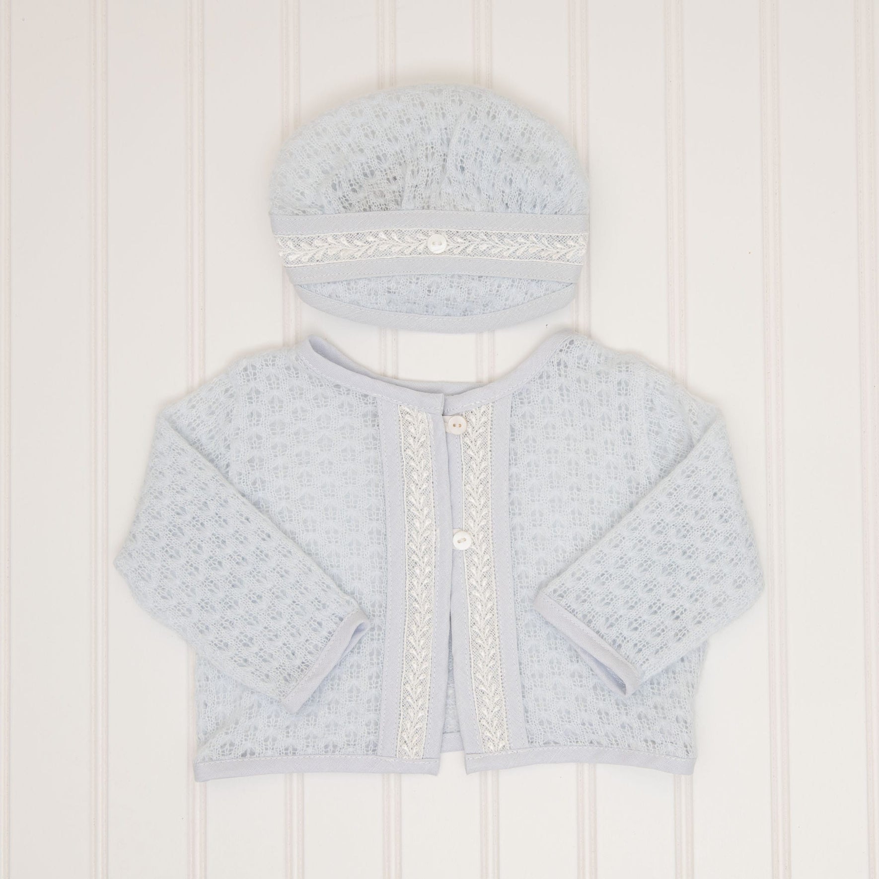 The Harrison Newborn Sweater, a soft blue knit cardigan featuring lace detailing and buttons, is displayed flat on a textured white surface. Above it, a matching hat adorned with similar lace detail complements the set perfectly for a newborn christening.