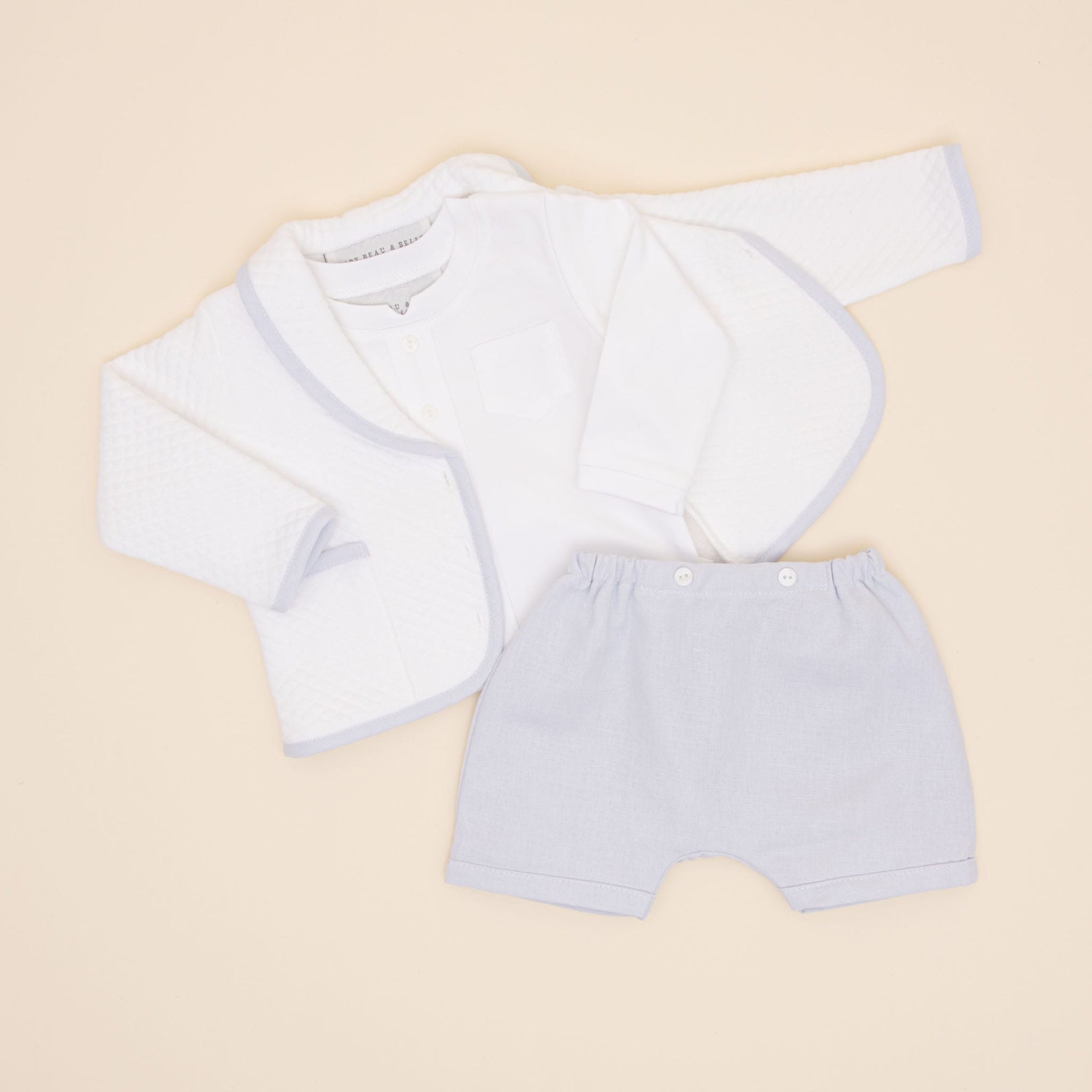 The Harrison Quilted Newborn Suit, a neatly arranged baptism outfit consisting of a white quilted jacket, a white long-sleeved shirt, and light gray shorts, is elegantly displayed on a light beige background.