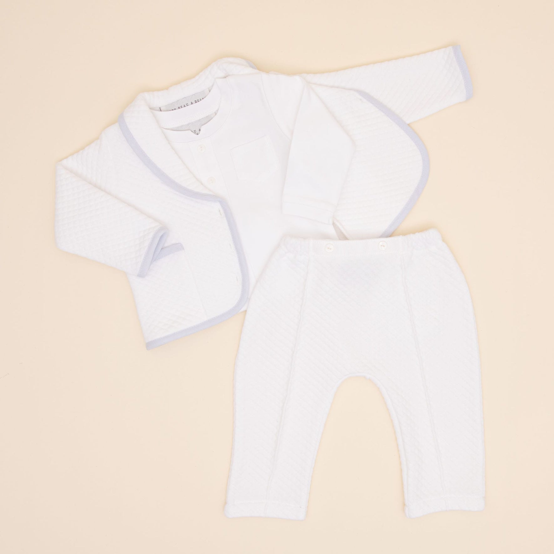 The Harrison Quilted Newborn Suit is beautifully displayed on a light surface, showcasing a quilted white jacket with subtle trim, a white top, and matching quilted pants. This adorable ensemble is perfect for newborn christening clothes, with each piece arranged to highlight its front.