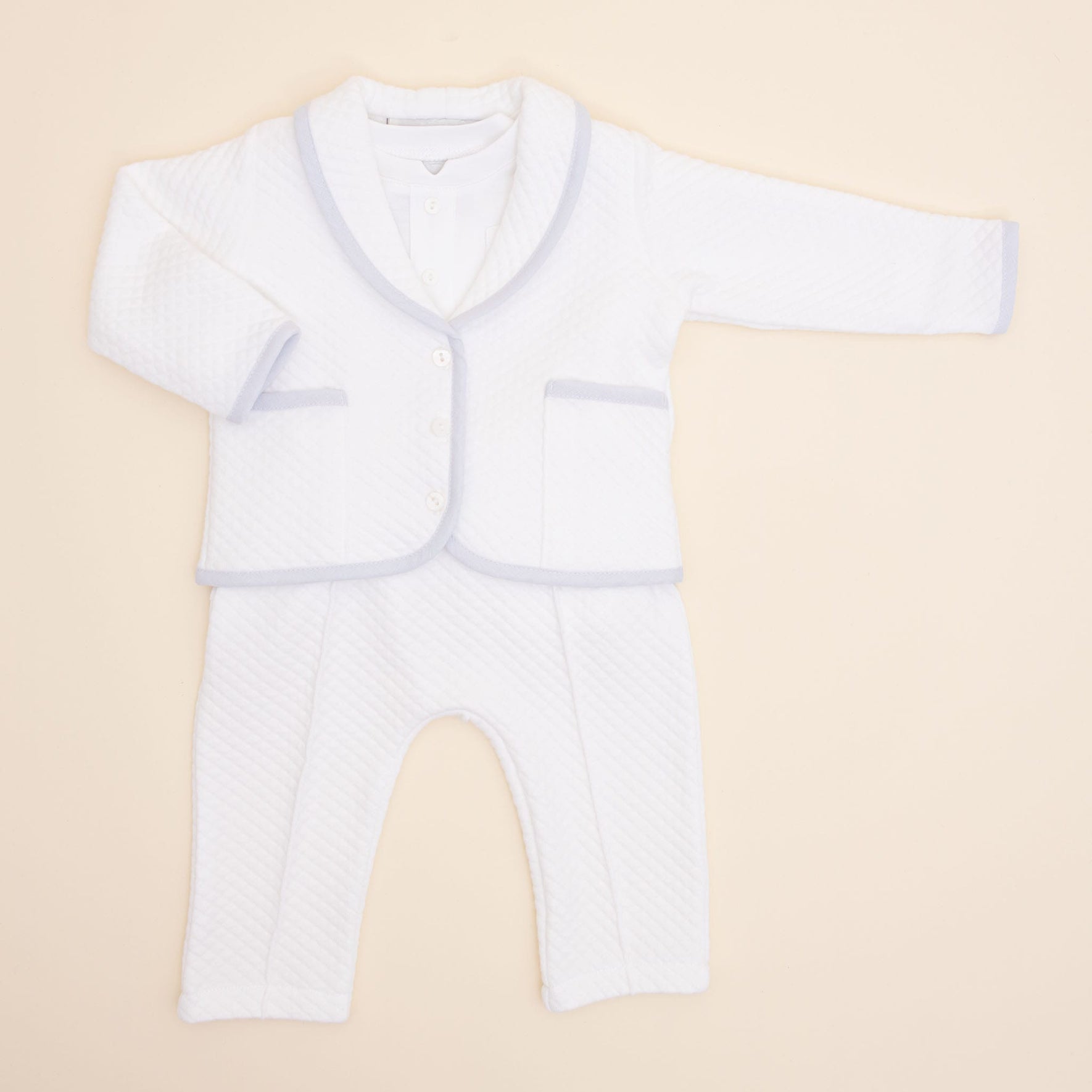 The Harrison Quilted Newborn Suit, an ideal choice for a baptism outfit, is displayed on a light beige background. This white ensemble features a long-sleeved jacket with buttons and matching pants. The quilted texture, complemented by light gray trim on the edges, adds an elegant touch suitable for special occasions.