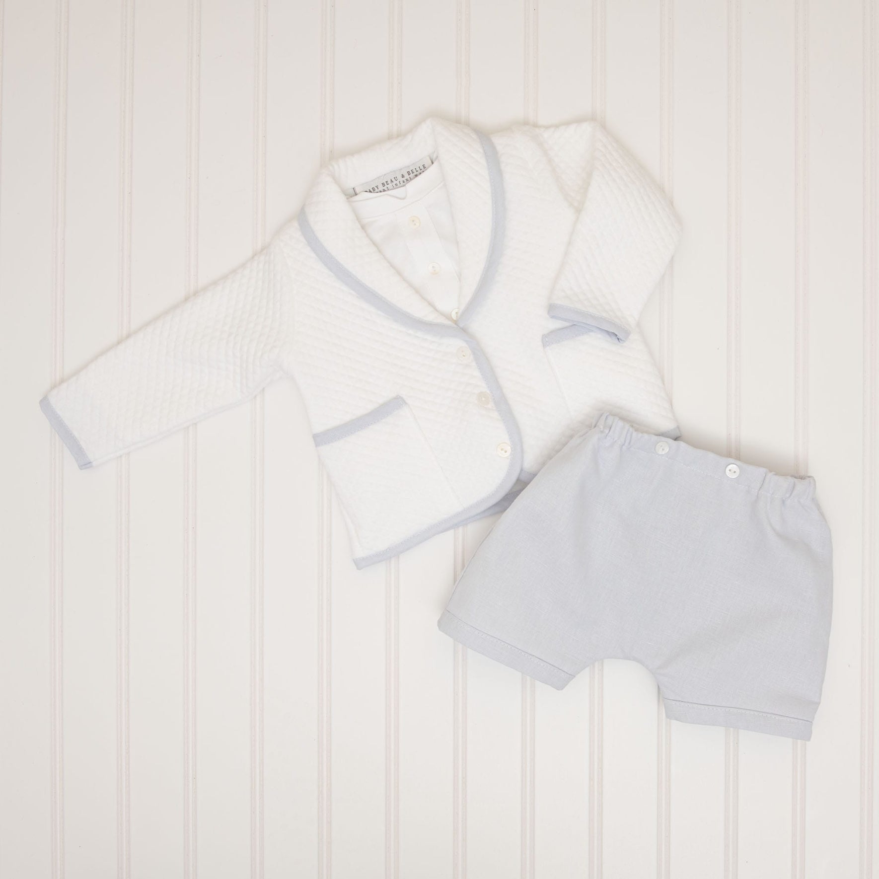 The Harrison Quilted Newborn Suit, ideal for a newborn christening, is showcased on a light background. It includes a white, textured cardigan accented with light blue trim and coordinating light blue shorts, all laid flat.