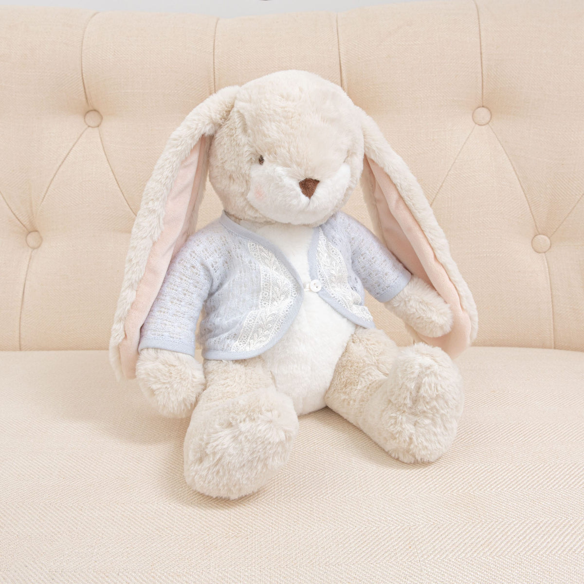 A Harrison Sweater Bunny from the Baby Safe Soft Plush collection sits on a light beige, tufted sofa. The bunny has long ears and wears a charming blue knit sweater, with soft cream-colored fur.