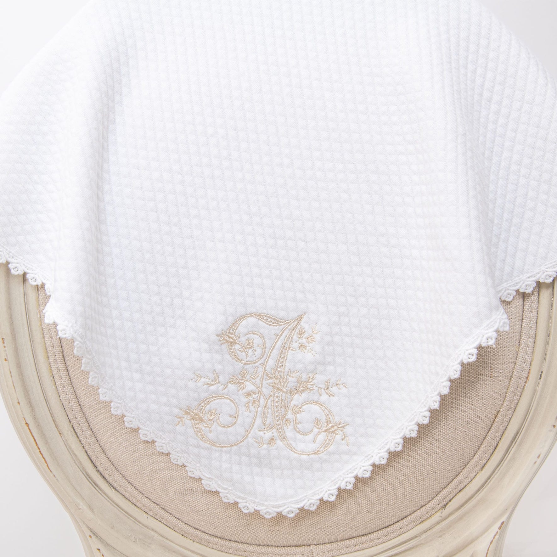The Initial Blanket is a white, quilted cotton piece with a waffle pattern that drapes over a curved chair back. It features personalized embroidery of an ornate letter "A" in beige near the bottom edge, complemented by a delicate lace trim along its lower boundary.