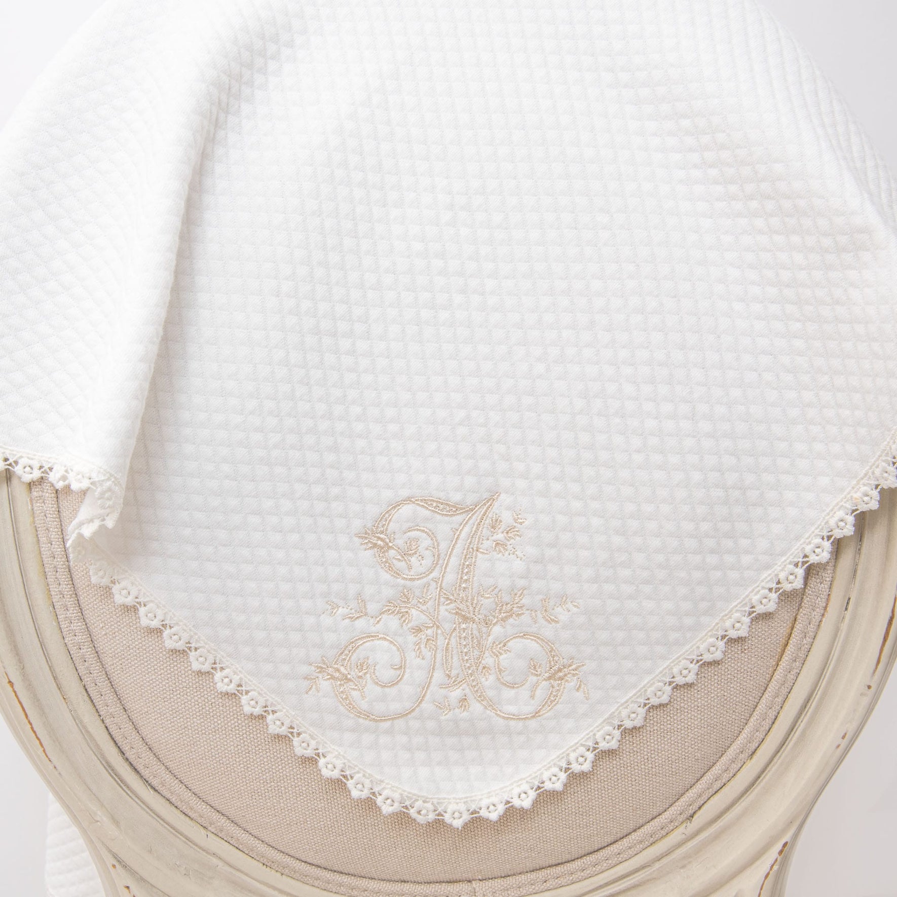 The Initial Blanket, a white quilted cotton baby blanket featuring a lace-trimmed edge, drapes elegantly over a beige upholstered chair. Adorned with an ornate embroidered letter "A" and light beige floral accents, it offers a charming touch of personalized embroidery.