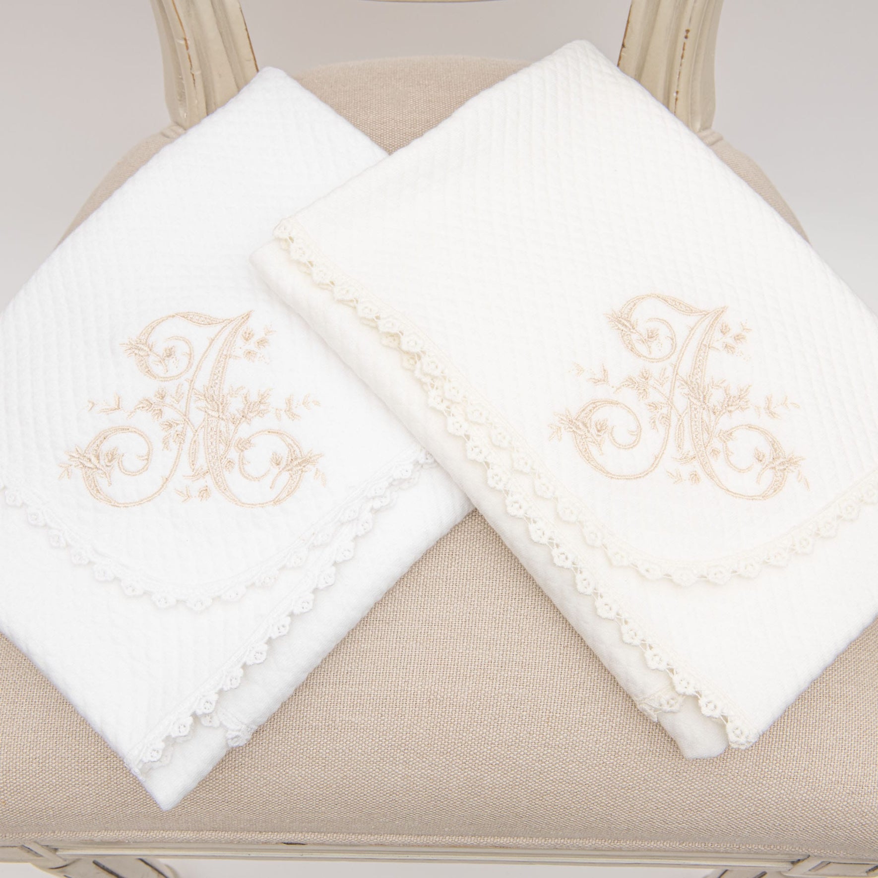Two folded white towels, featuring embroidered floral designs and lace trim, rest elegantly on a beige cushioned chair. Each towel displays monogrammed letters reminiscent of the delicate personalization found in the Initial Blanket.