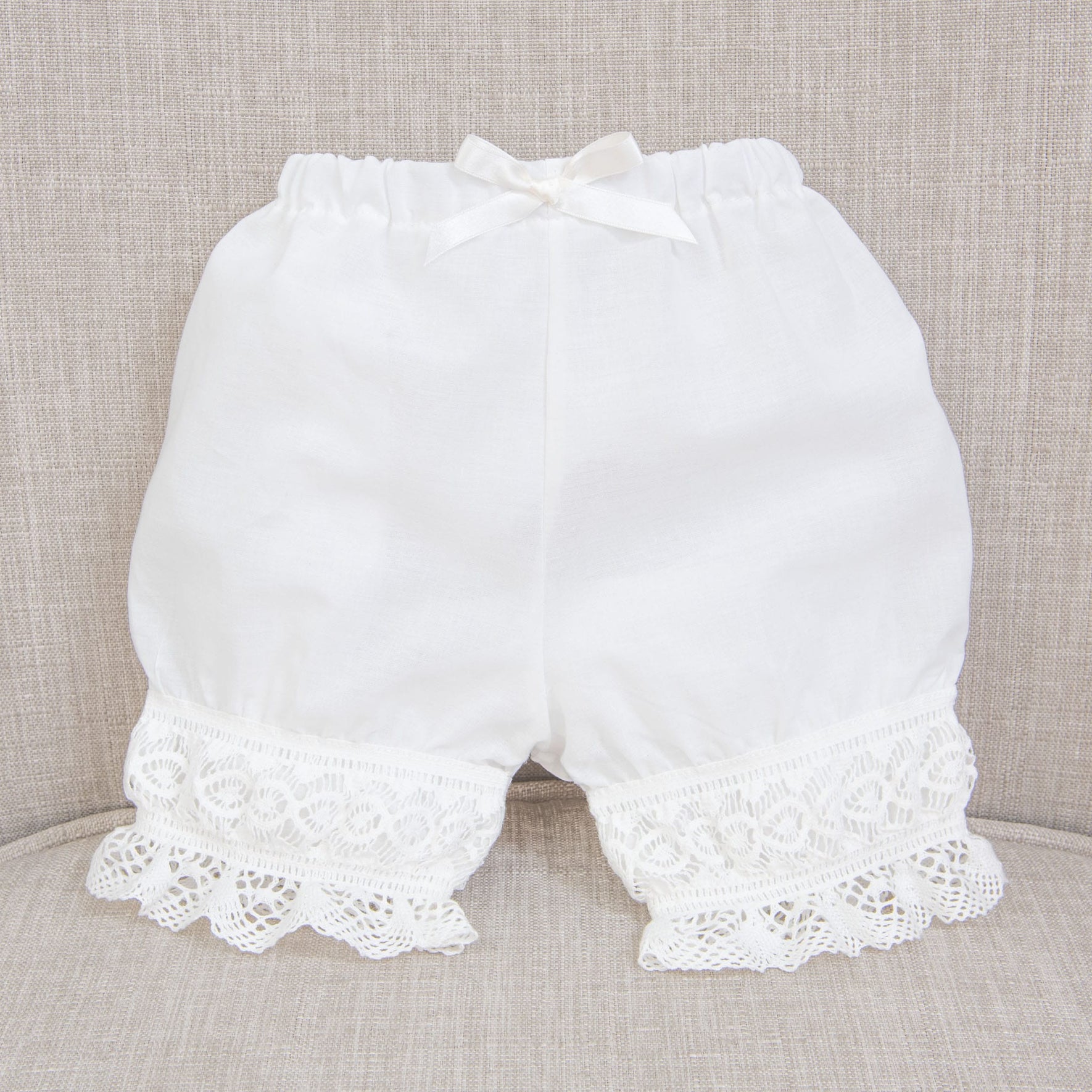 A pair of Isabella Bloomers, crafted from ivory cotton, features a delicate small bow at the waistband and charming lace trim around the leg openings. They are attractively displayed on a beige upholstered surface.