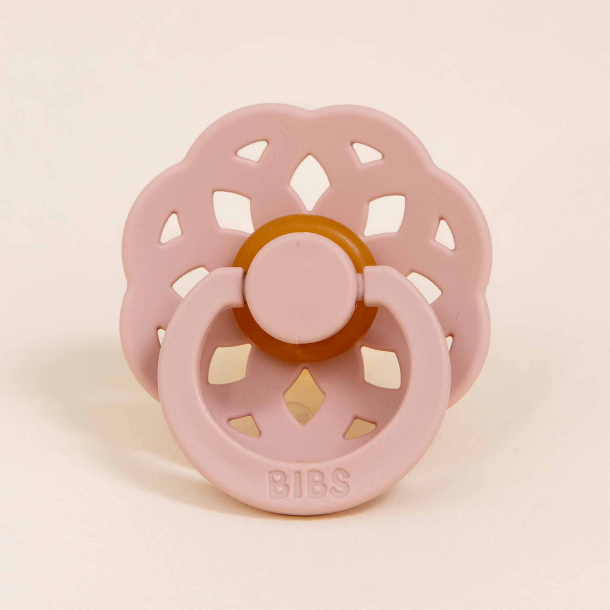 The Isabella Pacifier Set in Ivory & Blush features a pacifier with a flower-shaped outer shield, circular cutouts, and is crafted from natural rubber to aid in baby self-soothing. Its design includes a charming beige center, while the base of the nipple is embossed with "BIBS," highlighting its Danish origins.