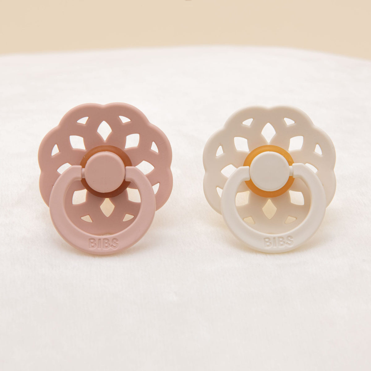 On a soft white surface, an Isabella Pacifier Set featuring two baby pacifiers with flower-shaped shields is beautifully displayed. One is in a blush shade, while the other is in ivory, both crafted from natural rubber designed to help babies self-soothe. The pacifiers proudly showcase the "BIBS" brand on their handles, highlighting this cherished choice from Denmark.