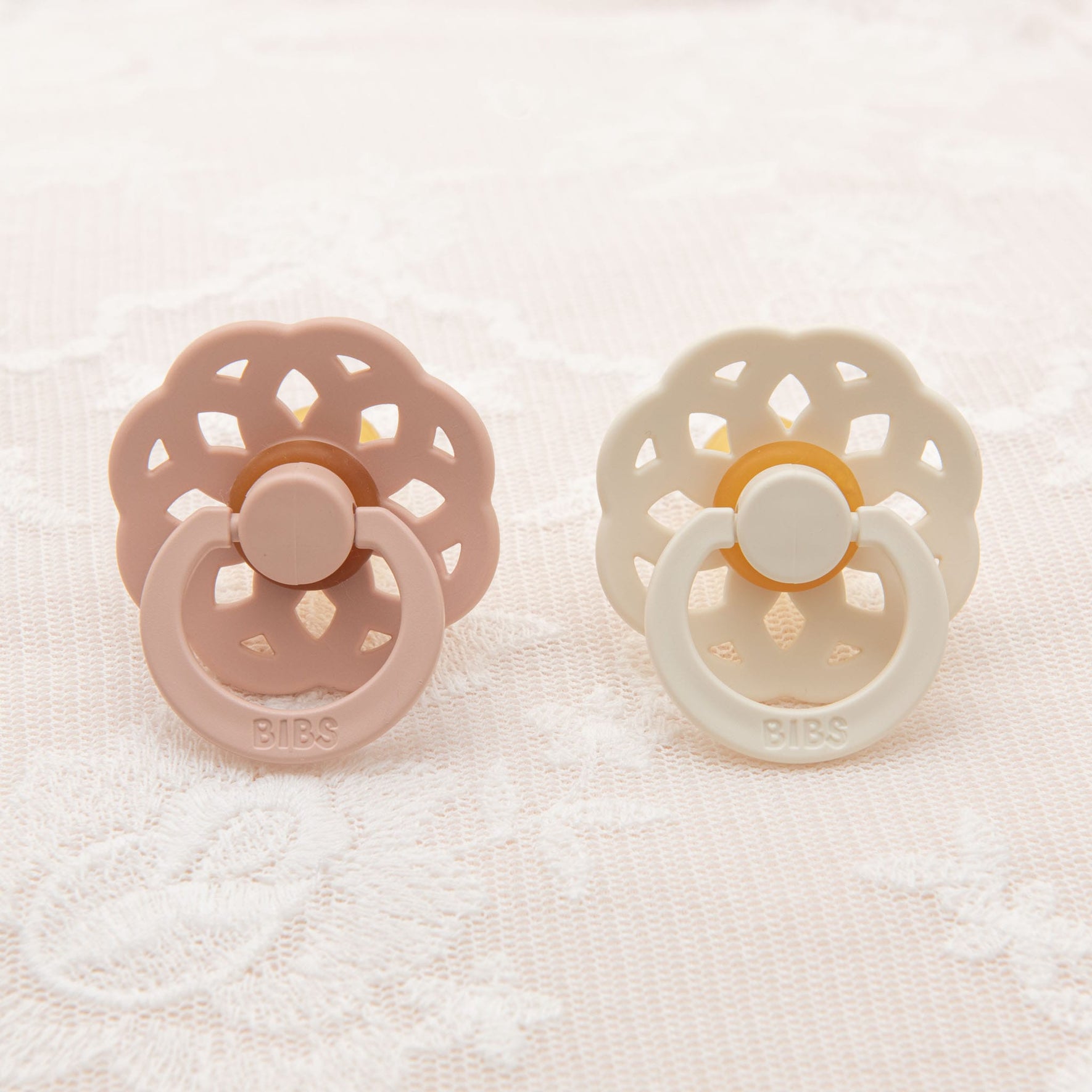 Two pacifiers from the Isabella Pacifier Set | Ivory & Blush with decorative floral backings rest on a lace-patterned surface. Crafted in Denmark, one blush-colored and the other ivory, each natural rubber baby pacifier features the word "BIBS" imprinted on its handle, perfect for helping little ones self-soothe.