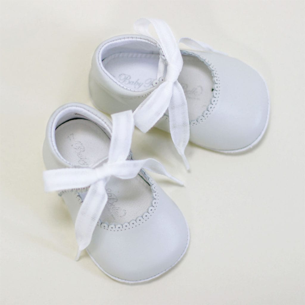A pair of Ivory Tie Mary Janes by Baby Beau & Belle, handmade with decorative white ribbons and a scalloped trim, gracefully rest on a plain light surface in an overhead shot.