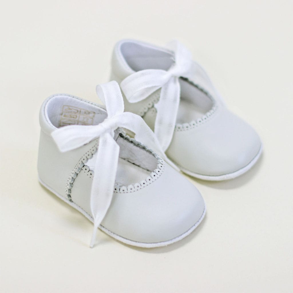 The Ivory Tie Mary Janes, crafted by Baby Beau & Belle, showcase scalloped edges and delicate white ribbon ties, elegantly placed on a light surface. These handmade baby shoes beautifully blend classic style with a touch of sophistication.