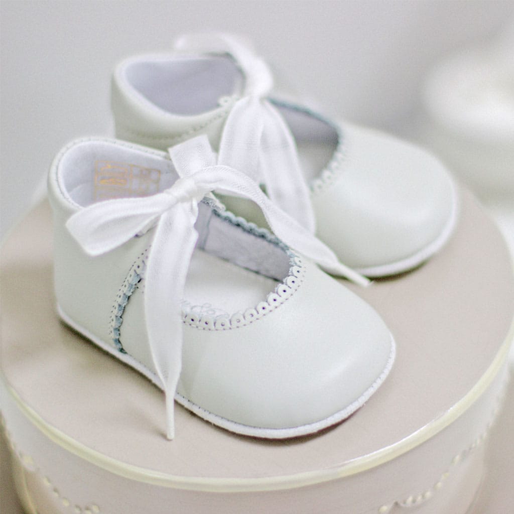 A pair of Baby Beau & Belle handmade baby shoes with ribbon ties rests on a light-colored circular surface, showcasing scalloped edges and a soft ivory sheen reminiscent of the Ivory Tie Mary Janes, all gently illuminated.
