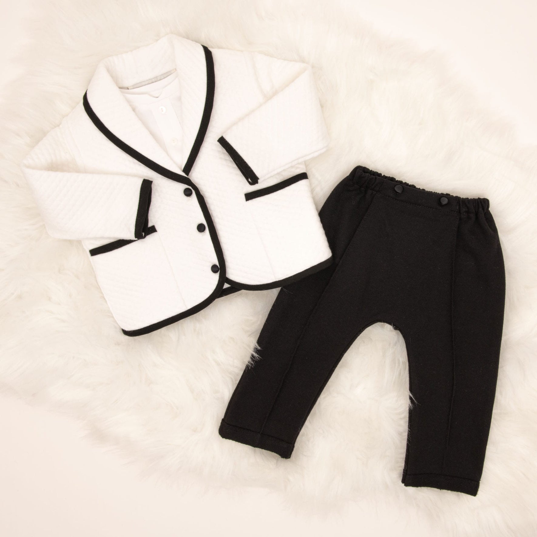 A flat lay of the James 3-Piece Suit, featuring the white quilted jacket with black trim and black pants, displayed on a soft fur background.