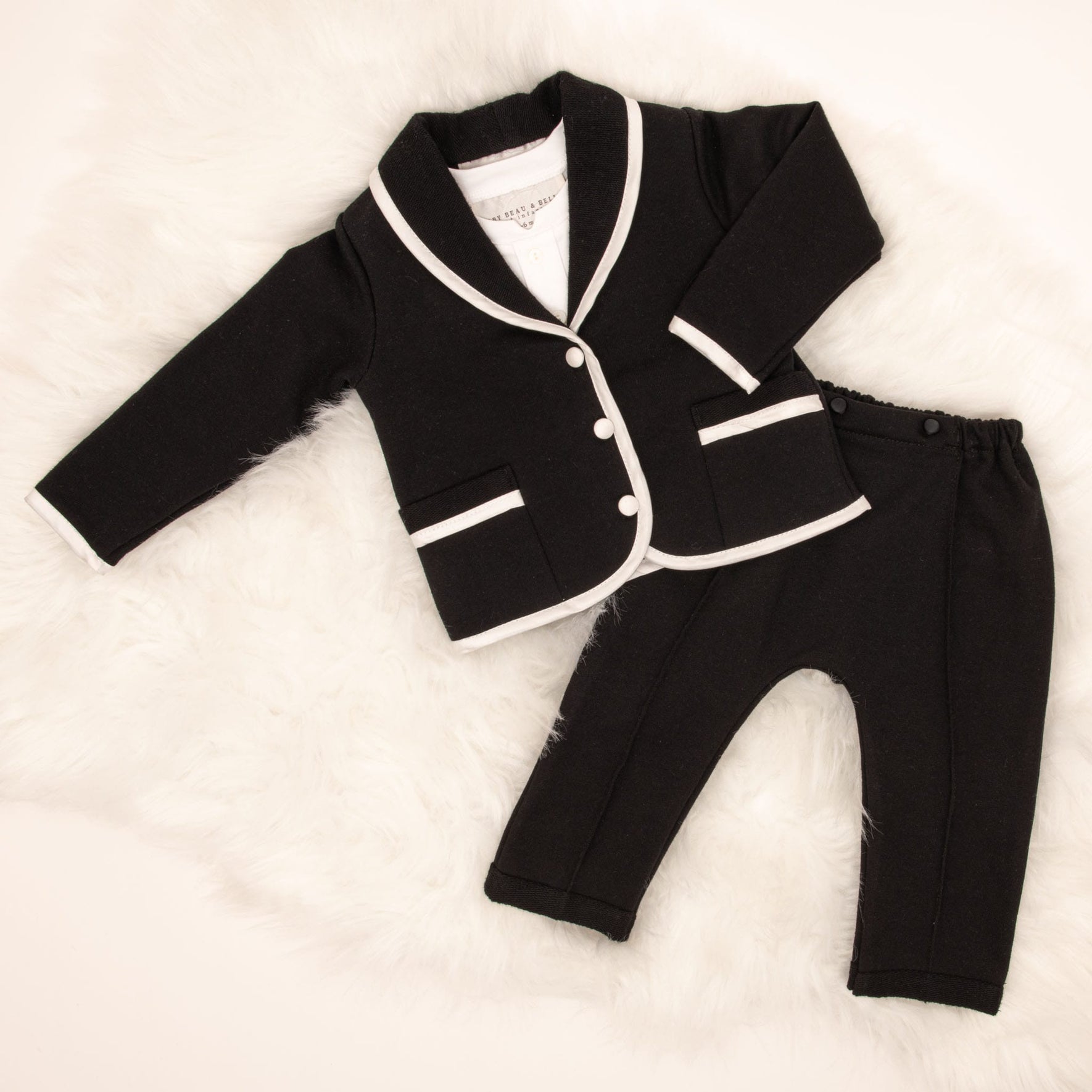 A flat lay of the James 3-Piece Suit in black, showing the neatly arranged black jacket with white trim and matching pants on a plush white surface.