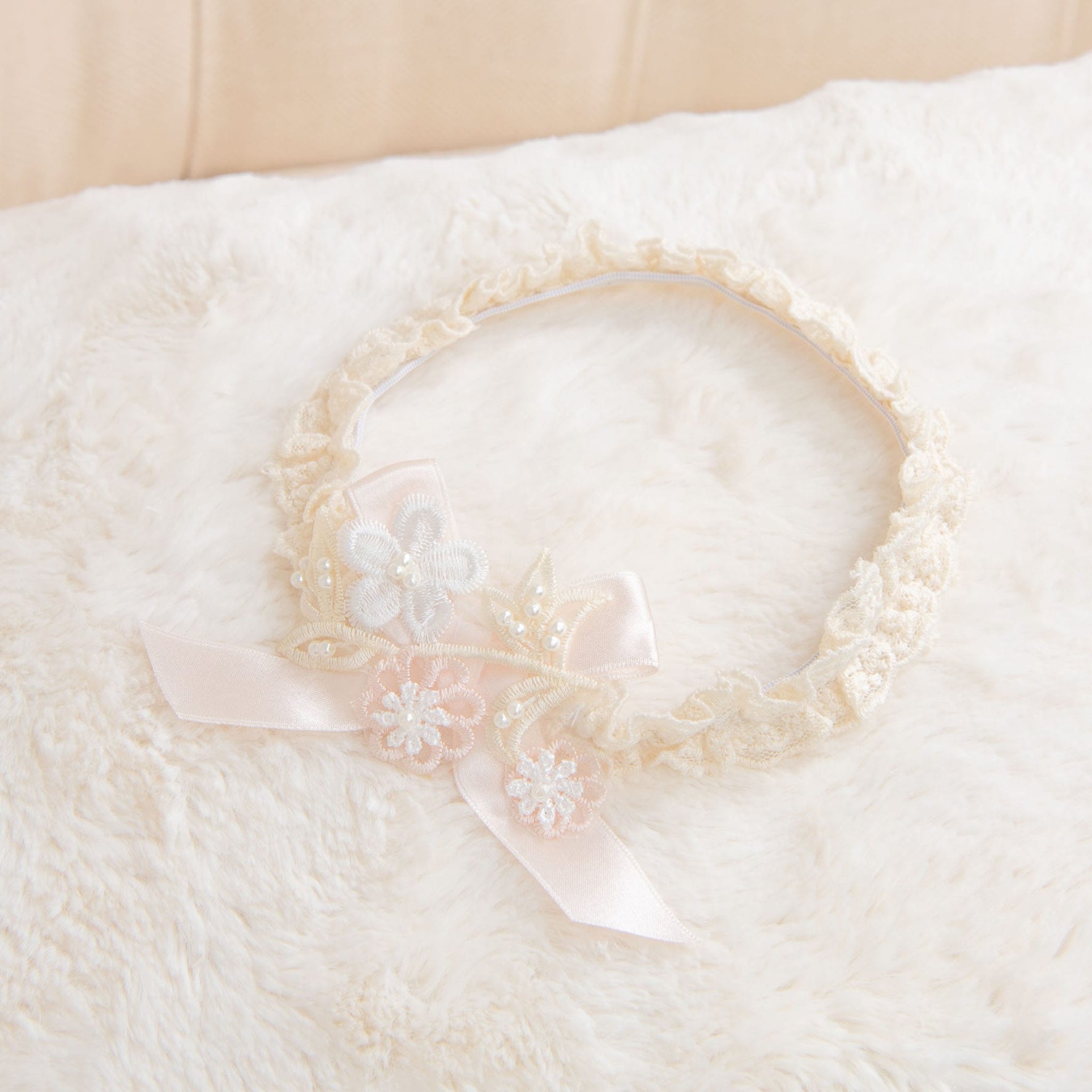 The Jessica Accessory Bundle, offering a 15% savings, includes a beige lace headband adorned with a pink ribbon and floral decorations. It rests perfectly on a fluffy cream-colored surface, making it an ideal complement to any baby dress accessory set.