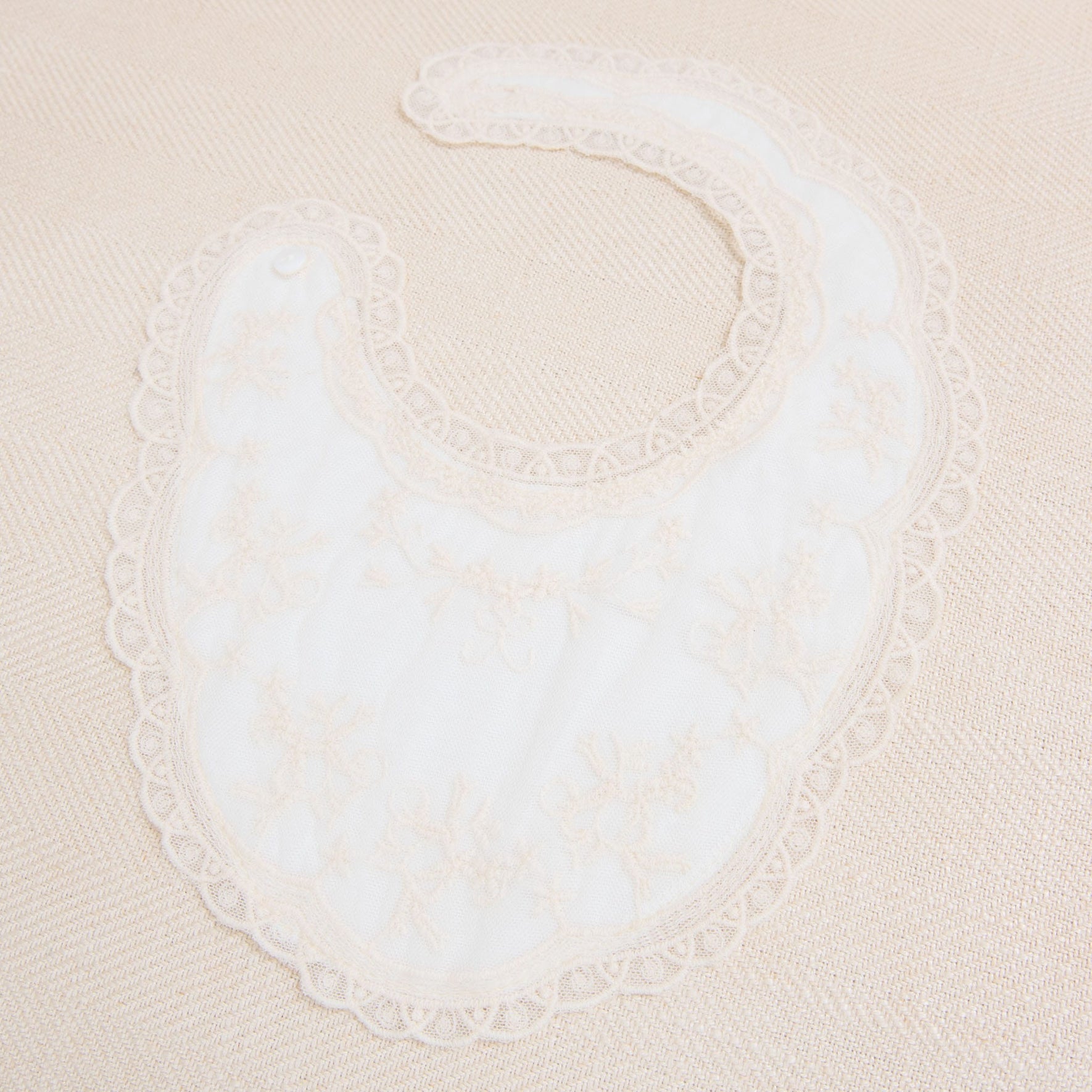 A Jessica Cotton Christening Bib, crafted from soft cotton, is laid out on a beige surface. The full-coverage bib features delicate floral embroidery and a scalloped champagne lace trim. Designed to fasten at the neck with a small snap button, this bib combines elegance with practicality.