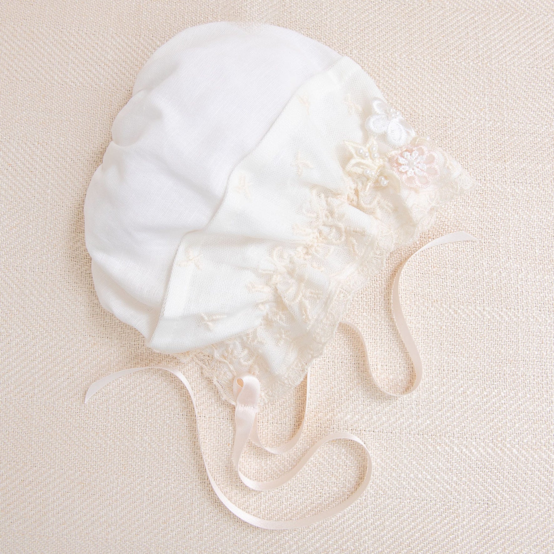 A white, vintage-style Jessica Linen Bonnet is adorned with champagne lace details and small embroidered flowers. It features a pair of soft, satin ribbons for tying and is displayed on a light, textured background.
