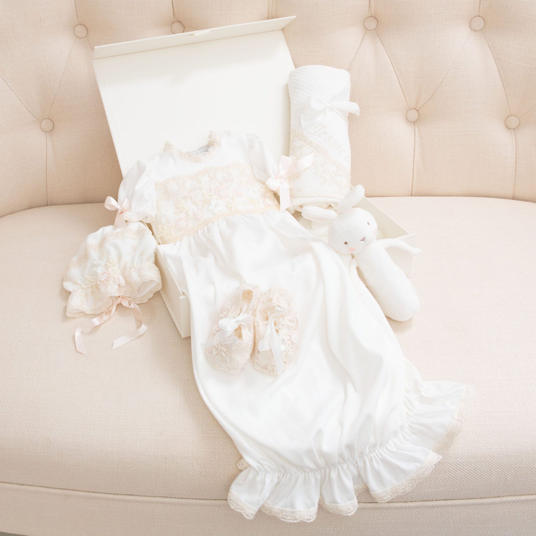 The Jessica Newborn Gift Set - Save 10% is neatly laid out on a light beige tufted sofa. The set, handmade in the USA, includes matching shoes, a bonnet, a blanket, and a white plush bunny toy. All items are placed next to an open, white gift box.