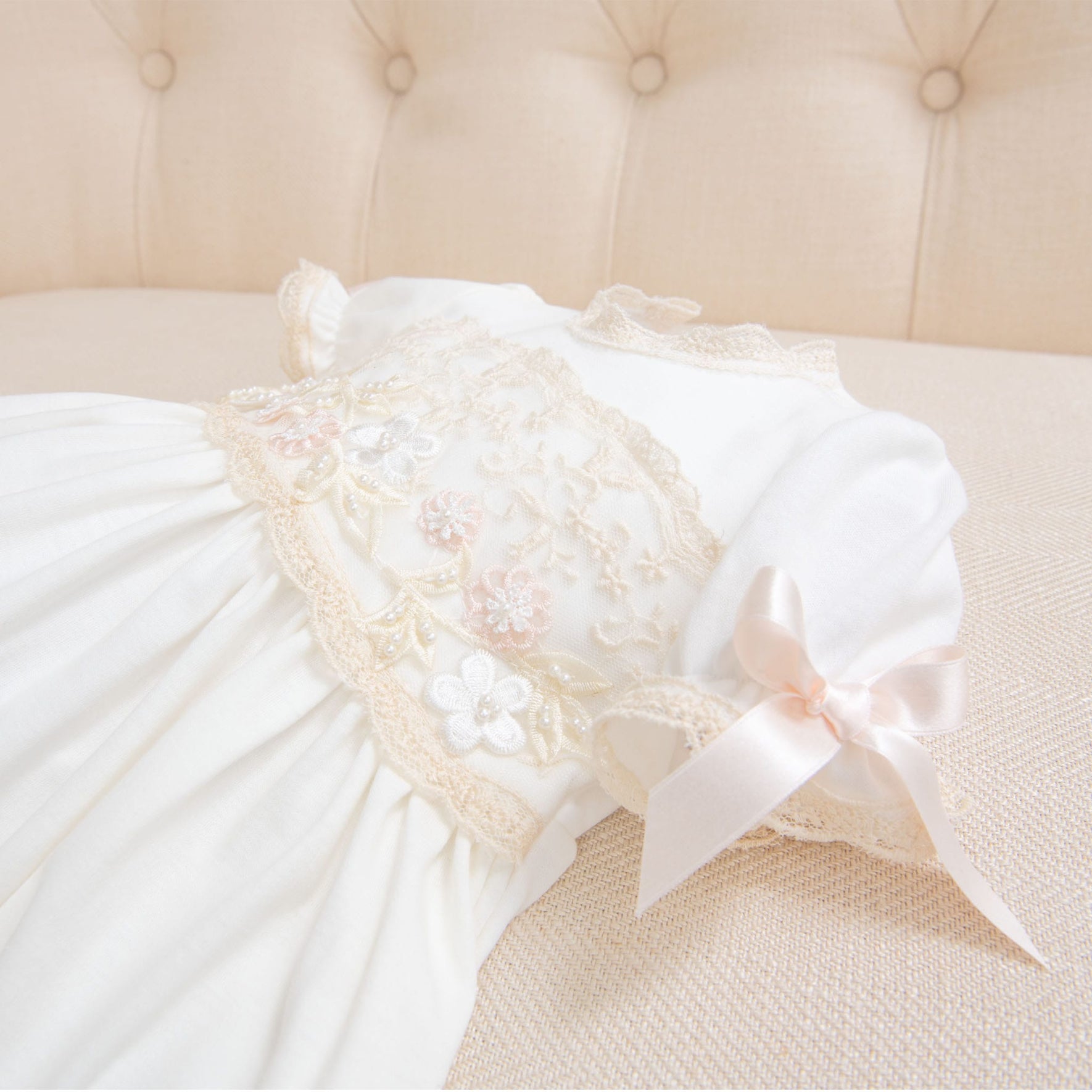 A close-up photograph of a Jessica Newborn Gift Set - Save 10% adorned with lace embroidery and small floral appliqués. The dress features short sleeves and a pink bow on one sleeve, perfect as a take me home outfit. It is placed on a beige, tufted background.