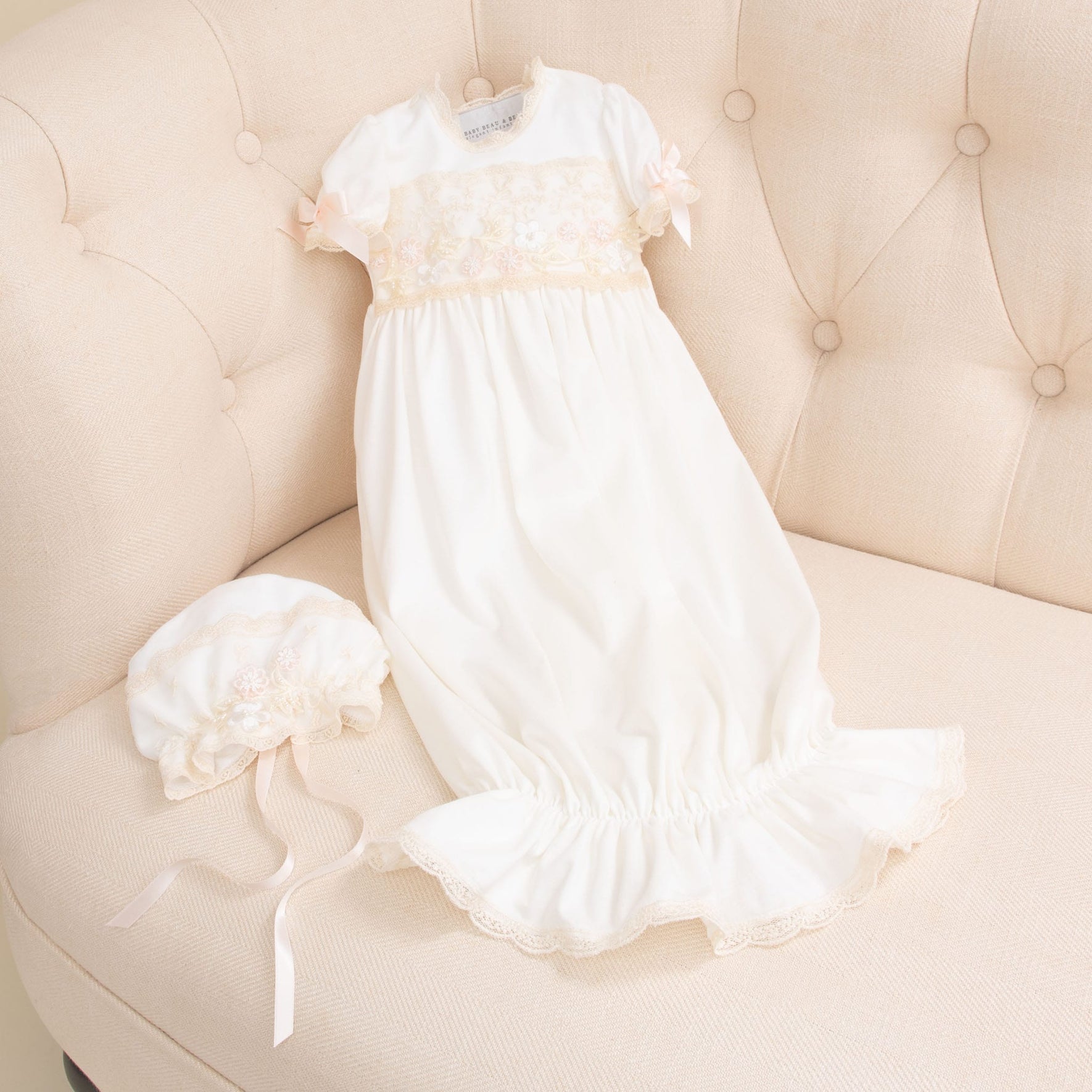 A Jessica Newborn Gift Set - Save 10% with lace detailing and a ruffled bottom is laid out on a cream-colored sofa. To the left of the set is a matching bonnet. Both items, handmade in the USA, feature delicate pink ribbon accents.