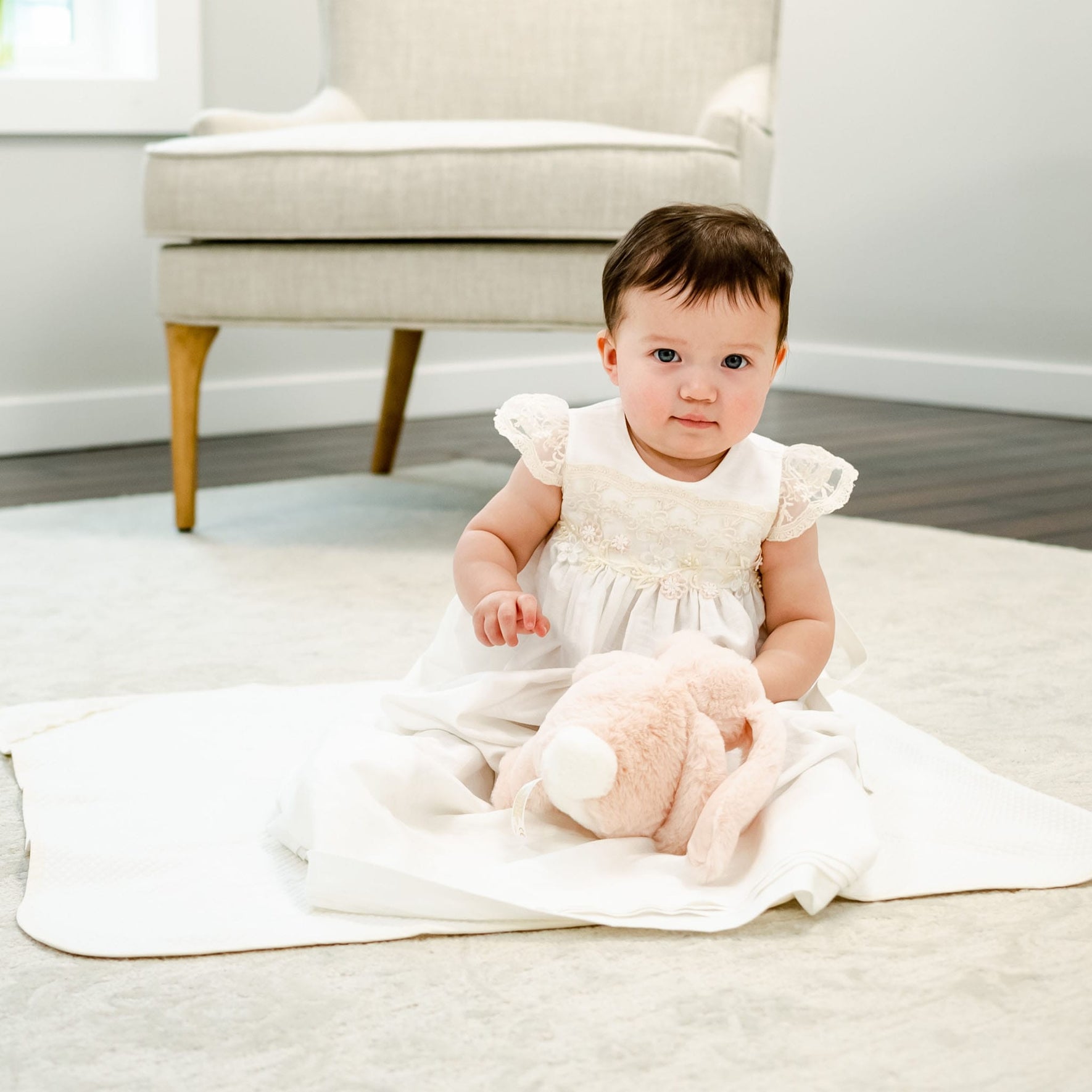 A toddler wearing a white dress is sitting on a Jessica Personalized Blanket on the floor. The child is holding a plush toy, perhaps a baby gift, and is in front of an upholstered armchair in a brightly lit room with wooden flooring.