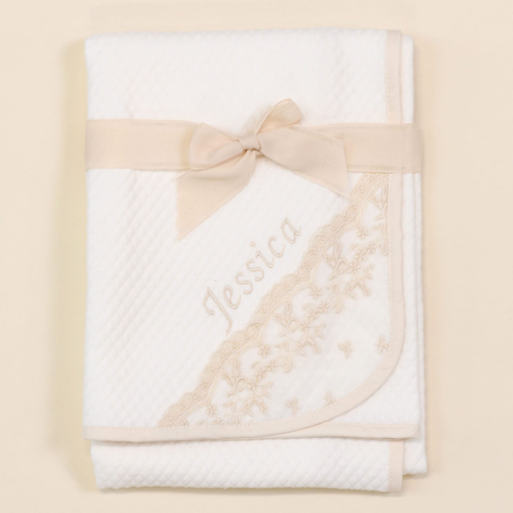 A folded white quilted *Jessica Personalized Blanket* with floral embroidery on the corner is placed on a light-colored surface. The handmade blanket is tied with a beige ribbon bow, and the name "Jessica" is embroidered in beige thread on the front, making it an ideal baby gift.