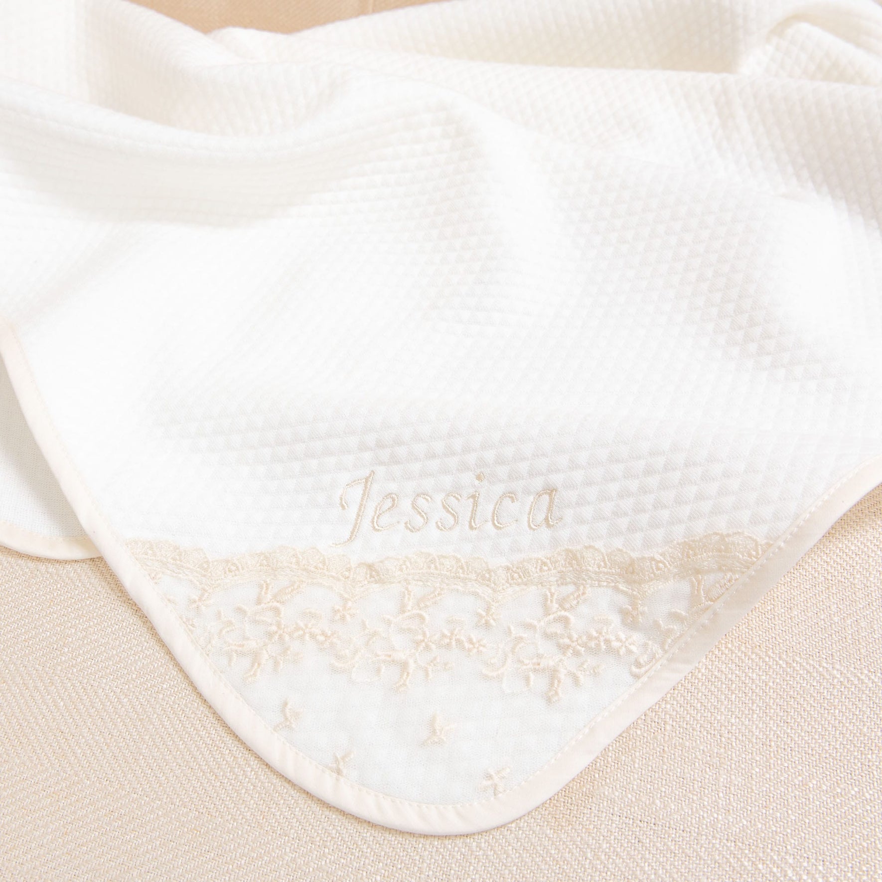 A white textured fabric, potentially a Jessica Personalized Blanket or towel, with an embroidered name "Jessica" in cursive near the edge. The bottom edge showcases an intricate floral lace pattern in cream. This handmade piece could make a charming baby gift against the soft beige background.