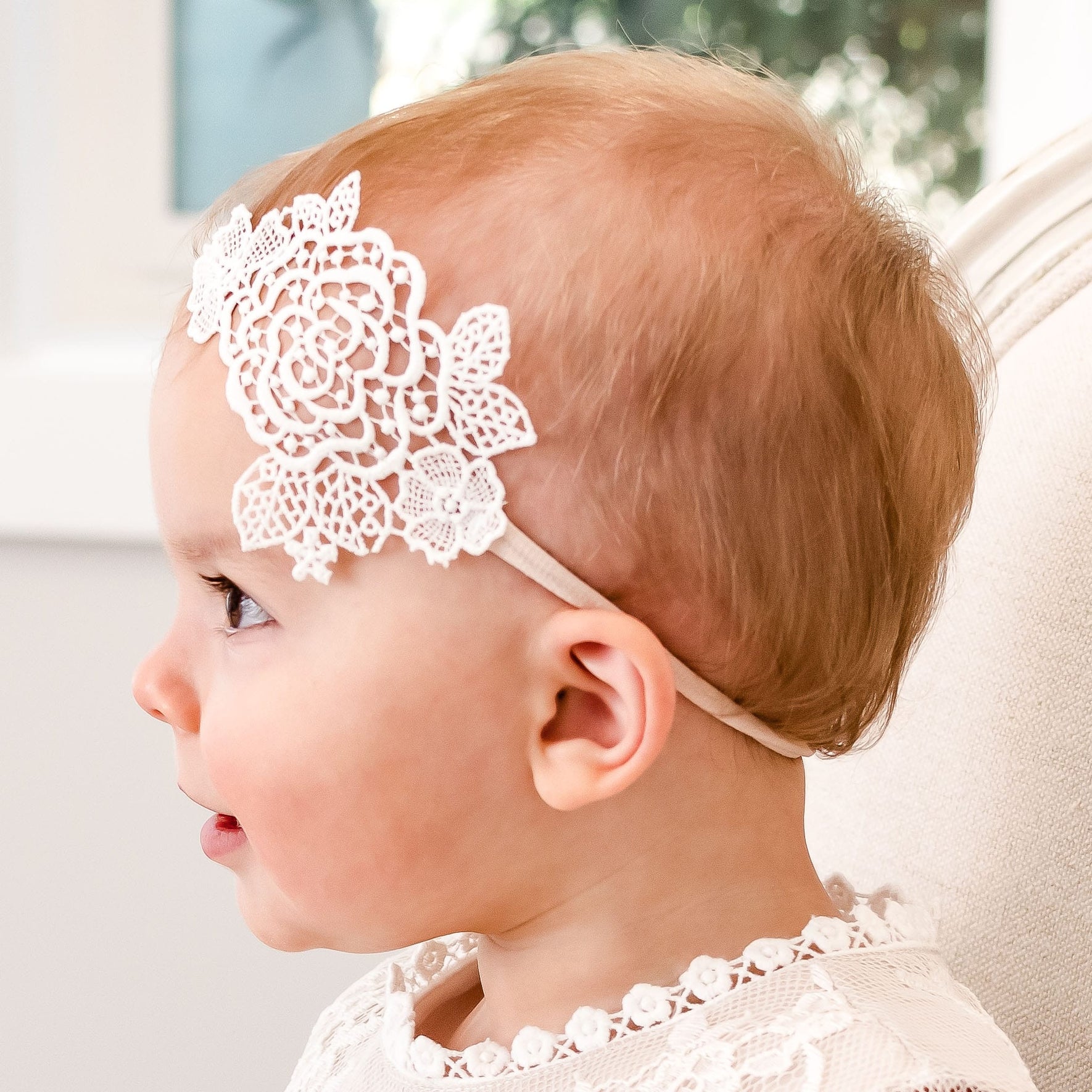 A baby wearing the Juliette Lace Headband, crafted from light ivory rose lace and hand-stretched nylon, is shown in profile. The baby has light brown hair and is dressed in a white outfit with a lace collar. The headband, handmade in the USA, adds an elegant floral touch to the ensemble. In the background, a white wall and a partially visible window can be seen.