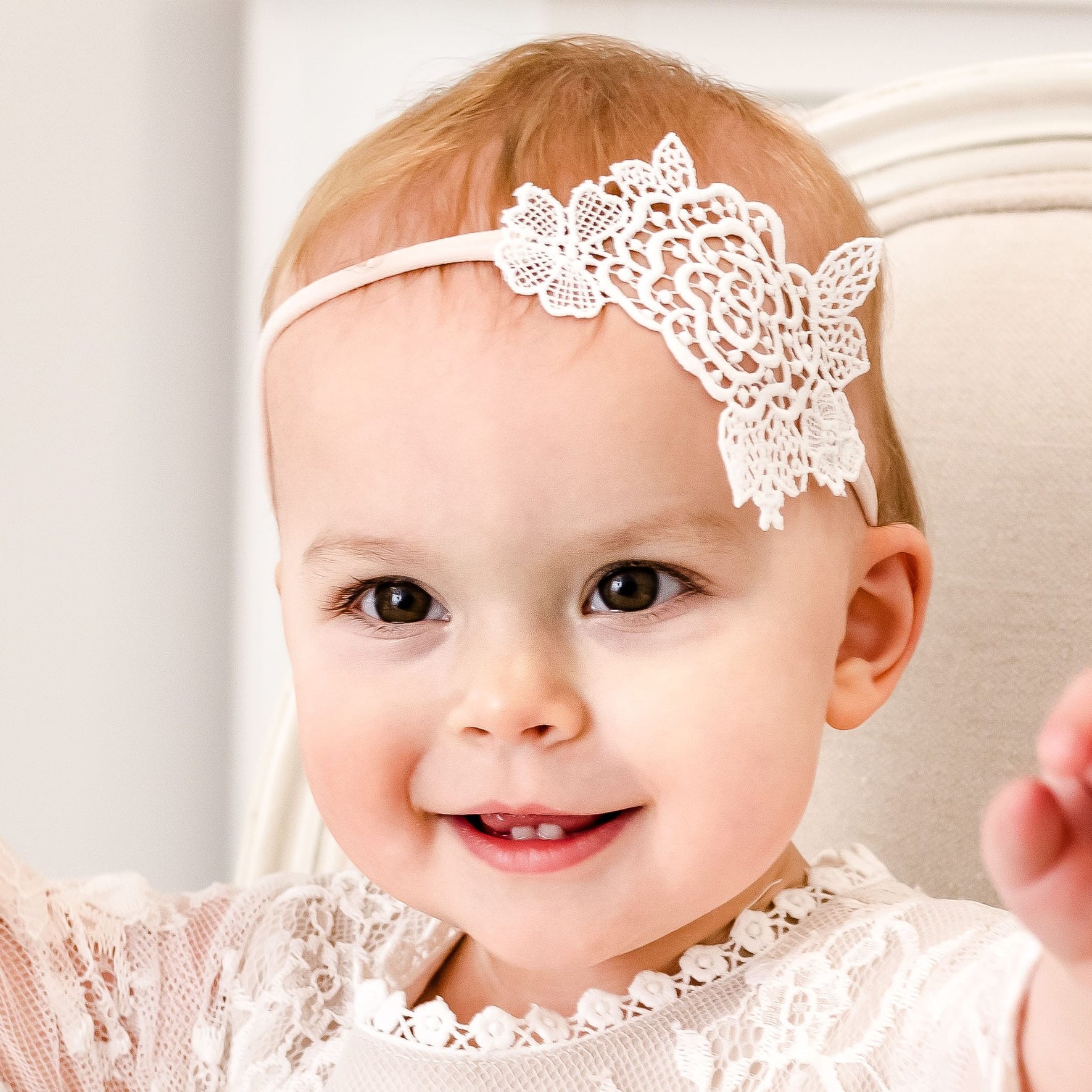A baby with light skin and short, light-colored hair is smiling. The baby is adorned in the Juliette Lace Headband featuring a decorative flower on the left side and dressed in an elegant light ivory rose lace outfit, handmade in the USA. They are sitting on a light-colored chair, radiating pure joy.