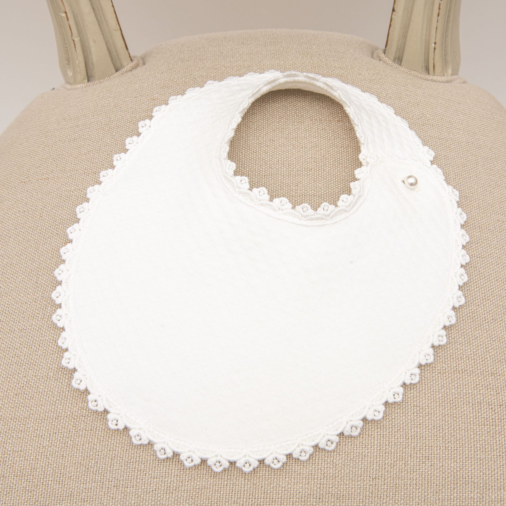 A close-up image of a baby bib with scalloped edges and lace detailing, displayed on a beige chair.