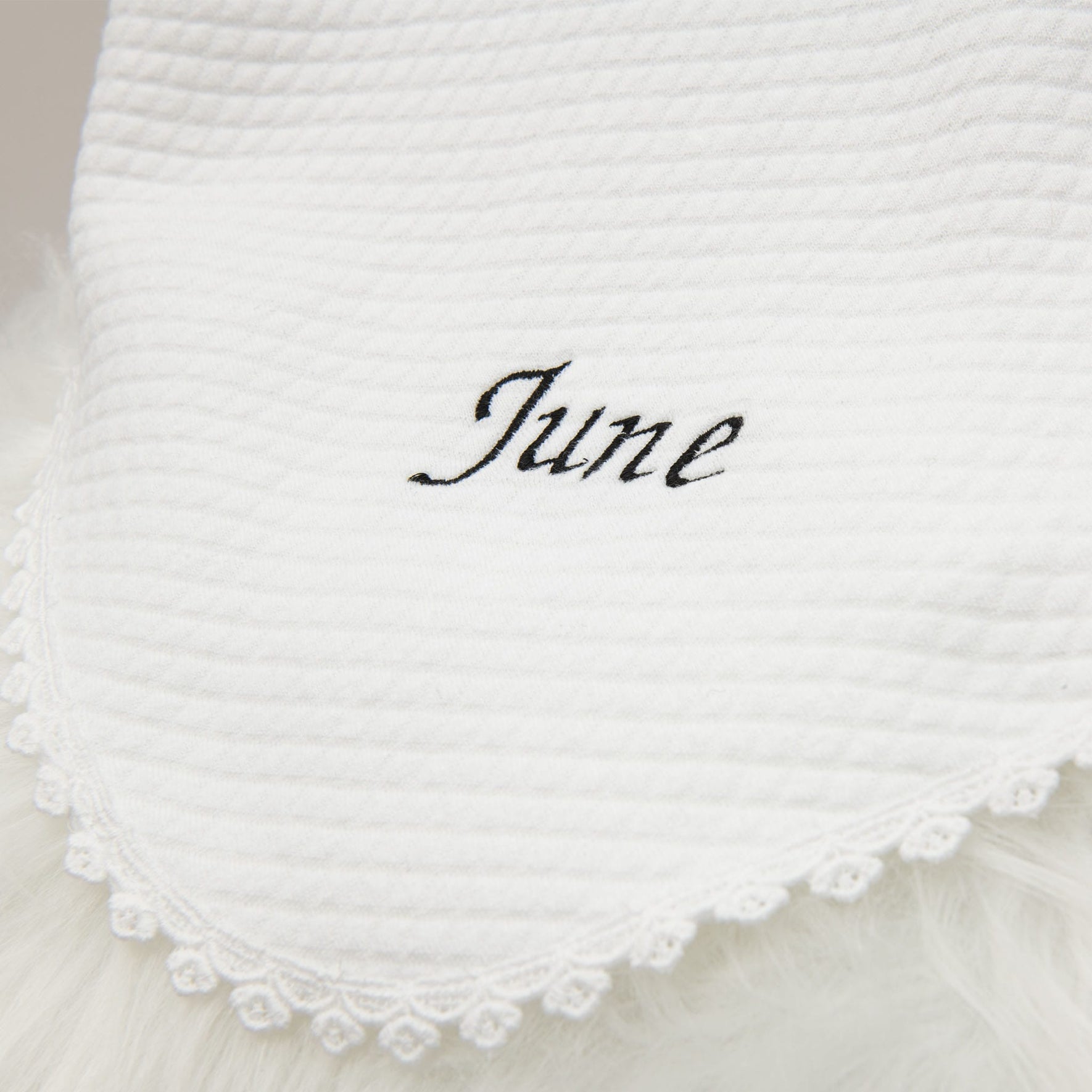 Close-up of a stylish white baby blanket with the name June embroidered in elegant black script, accented by a detailed lace trim along the edge.