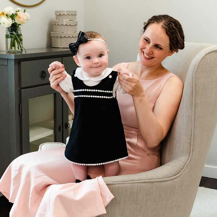 A woman in a stylish Dress Set sits in a gray armchair, holding a baby girl dressed in a black and white outfit after her christening. Both are smiling in a bright, cozy room.