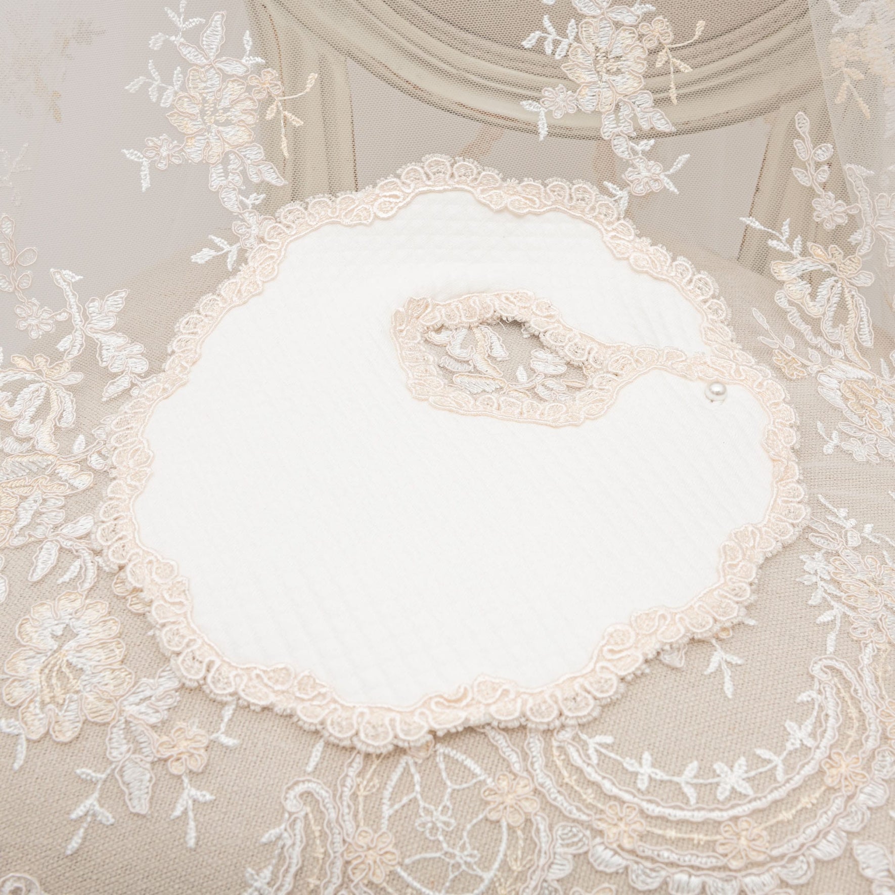 Introducing the Kristina Accessory Bundle - Save 15%: a beautifully crafted white bib with scalloped edges and delicate lace trim, elegantly showcased on a beige lace surface adorned with floral embroidery. This bib is the perfect accessory to enhance any baby girl's Christening ensemble, featuring a convenient snap button closure for effortless wear.