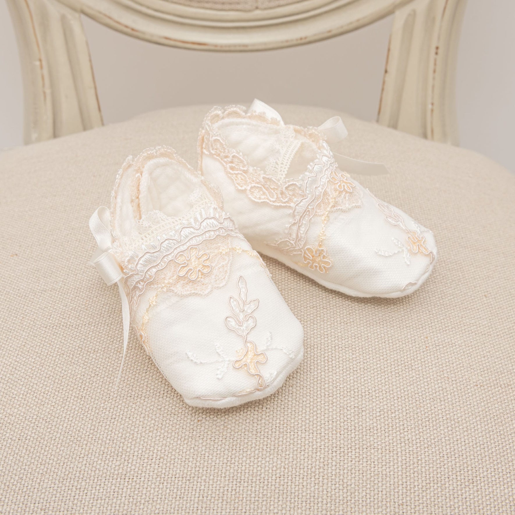 Two white baby shoes from the Kristina Accessory Bundle, featuring delicate lace and floral embroidery, rest on a beige fabric surface. Ideal for a baby girl Christening outfit, these shoes showcase small ribbon bows and intricate detailing, offering an elegant appearance. Get them now with a 15% savings!