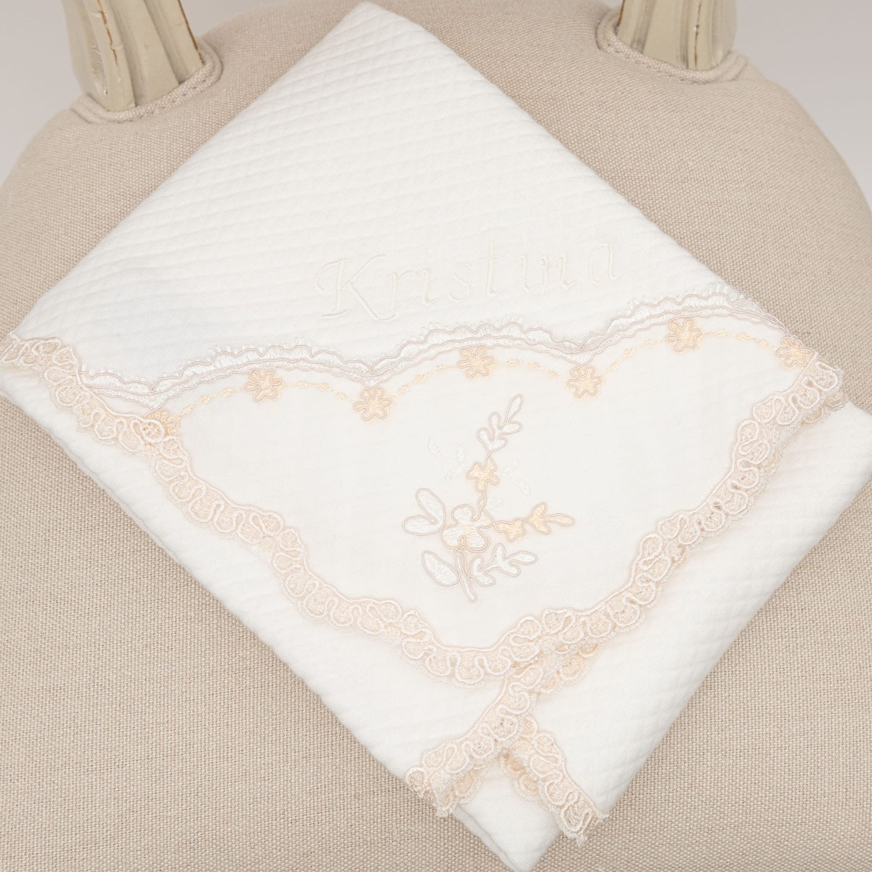 A folded white accessory from the Kristina Accessory Bundle, featuring delicate beige lace trim and an embroidered floral design, lies on a textured beige surface, reminiscent of a baby girl Christening outfit. The name "Kristina" is subtly displayed on the elegant fabric.