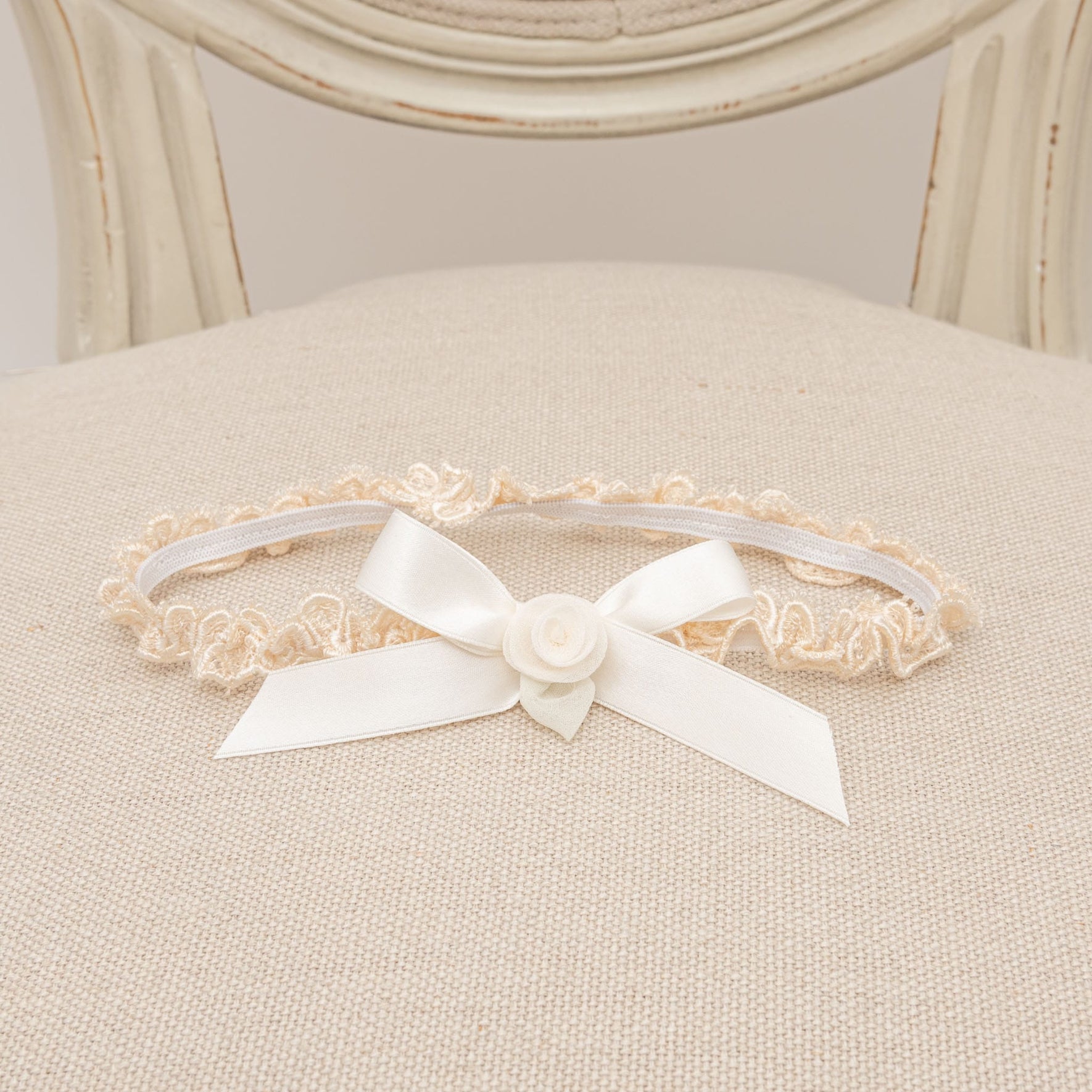 The Kristina Accessory Bundle, which saves you 15%, includes an elegant beige lace garter adorned with a white satin bow and rosette, beautifully enhancing the delicate charm of a baby girl Christening outfit as it graces a beige cushioned chair.
