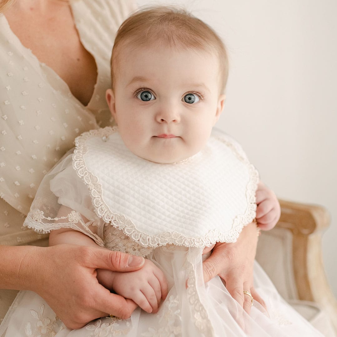 A baby with large eyes, wearing a delicate christening gown adorned with Venice lace and the Kristina Bib, is being held by an adult. The adult is in a light-colored dress, and both are seated against a neutral background.