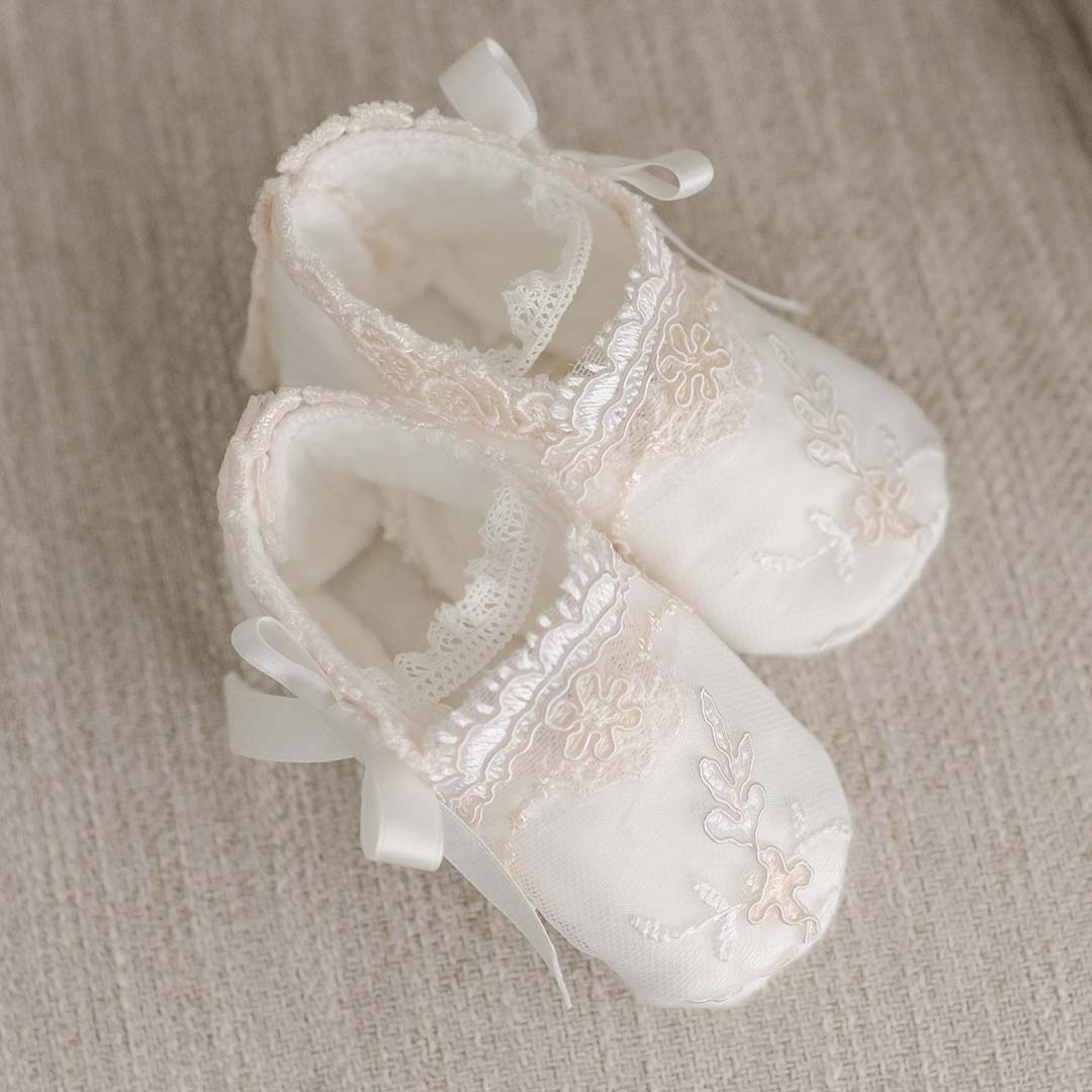 A pair of Kristina Booties, featuring delicate lace and floral embroidery accents, are placed on a textured beige surface. Small silk ribbon bows adorn the sides, perfectly complementing a christening gown.