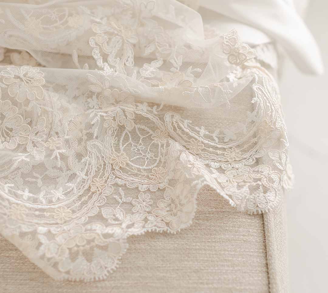 Close-up of white lace fabric with floral embroidery draped over beige textured material. The lace is delicate and intricate, comparable to the elegant finesse featured in the Kristina Christening Gown & Bonnet, showcasing detailed work against the solid background.