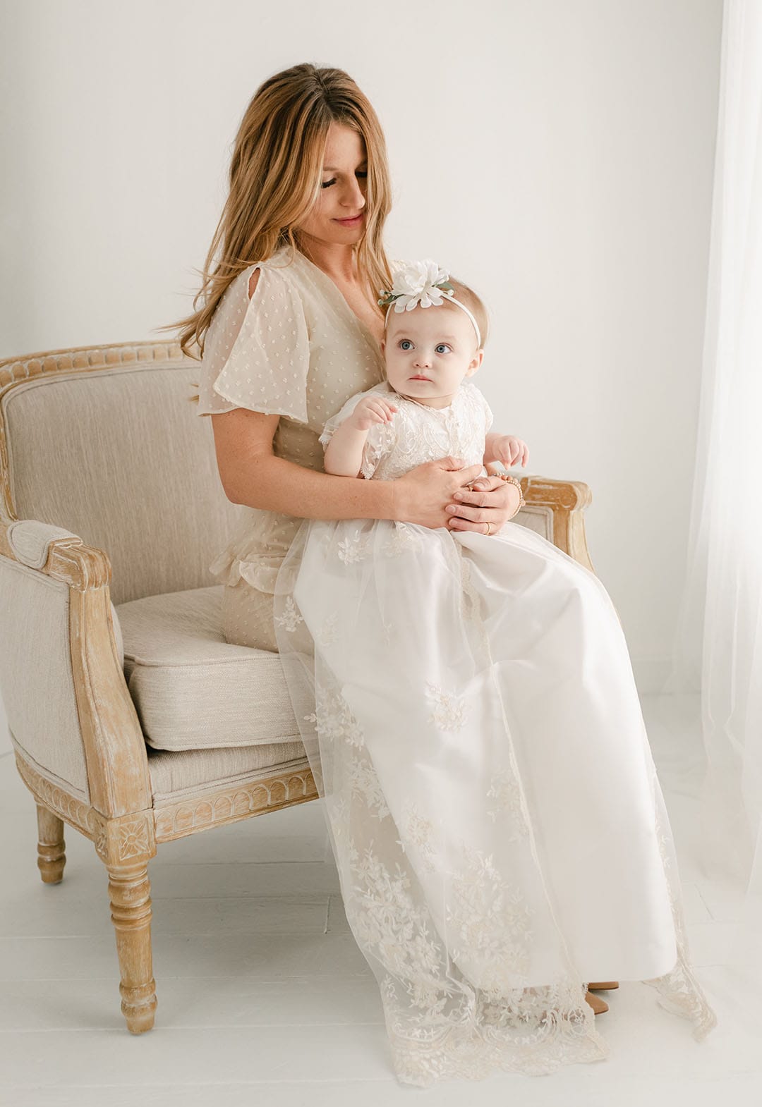 A woman is seated on a beige, vintage-style chair, holding a baby dressed in the Kristina Christening Gown & Bonnet, which features delicate lace and floral embroidery. The woman is wearing a light, sheer dress as they pose in a softly lit, neutral-toned room that exudes timeless elegance.