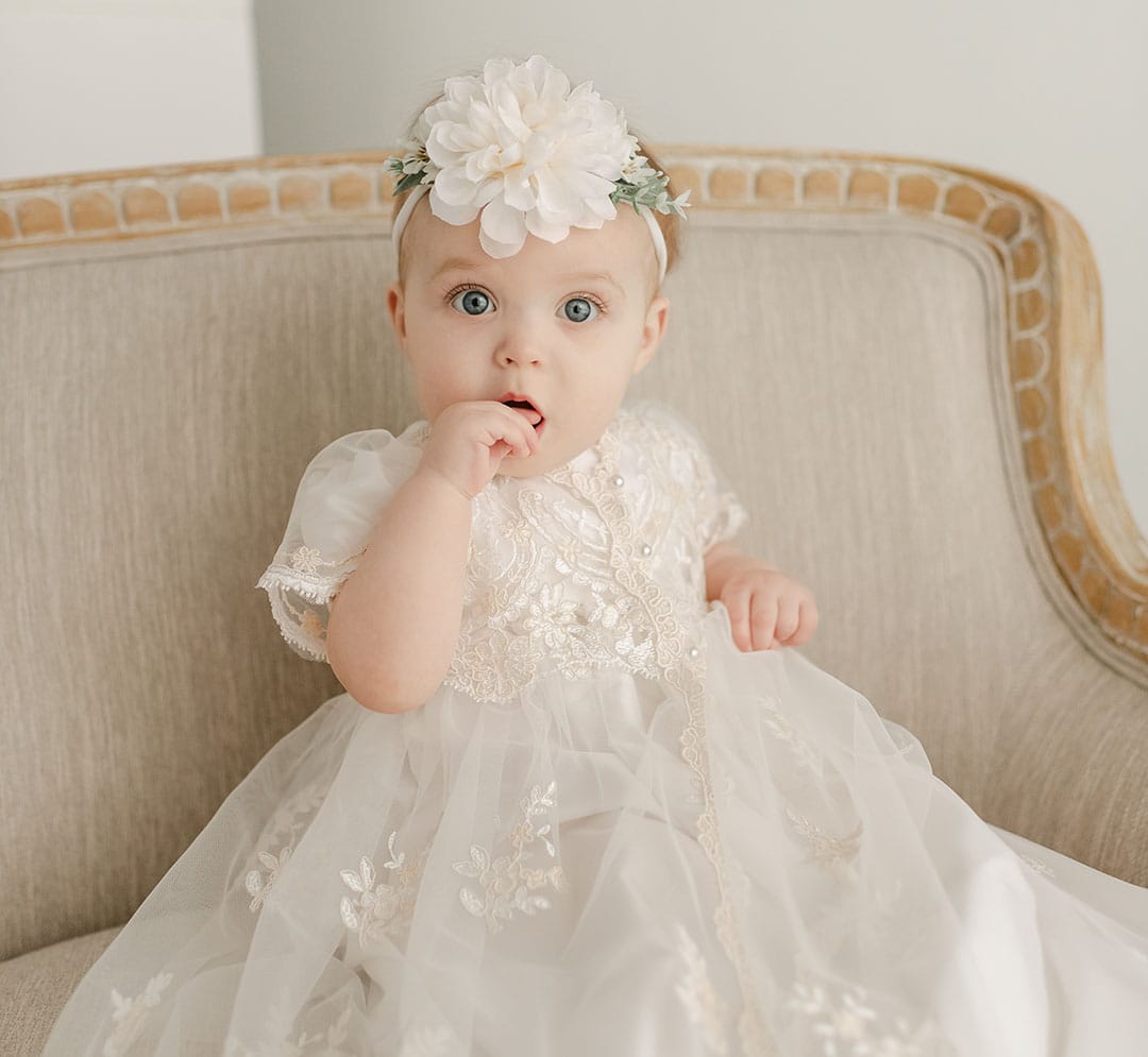 A baby, dressed in a white Kristina Christening Gown with an accompanying bonnet and adorned with a floral headband, sits on a beige upholstered chair. The baby's wide eyes sparkle as they touch their mouth with one hand.