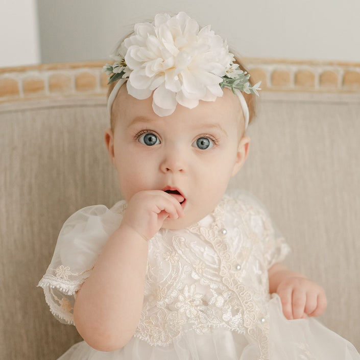 A baby with big blue eyes, wearing a white dress and the Kristina Flower Headband adorned with an ivory dahlia, sits on a beige chair. The baby has one finger in its mouth, gazing towards the camera.