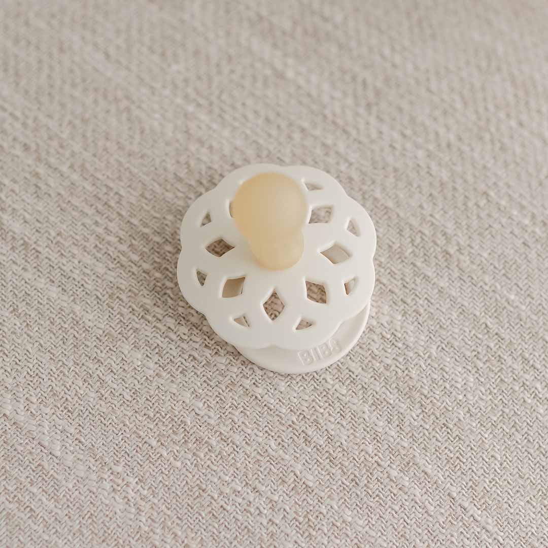 A silicone pacifier from the Kristina Pacifier Set in Vanilla & Ivory, featuring a honeycomb-patterned shield that resembles natural rubber, rests on a textured beige surface. Ideal for little ones to self-soothe, this elegantly designed item comes from Denmark.